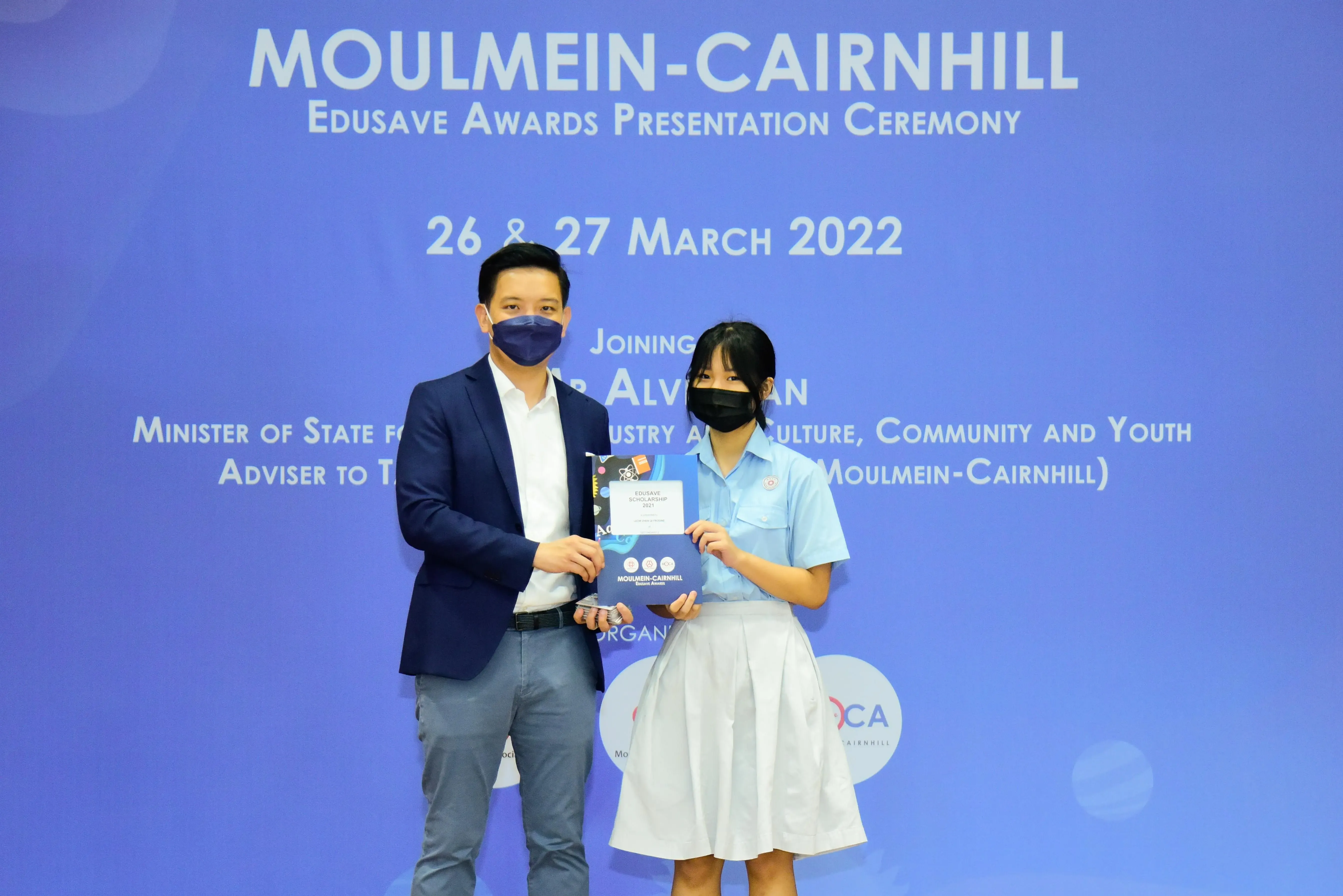 Two masked individuals hold an award booklet at the Moulmein-Cairnhill Edusave Awards Presentation Ceremony, March 2022.