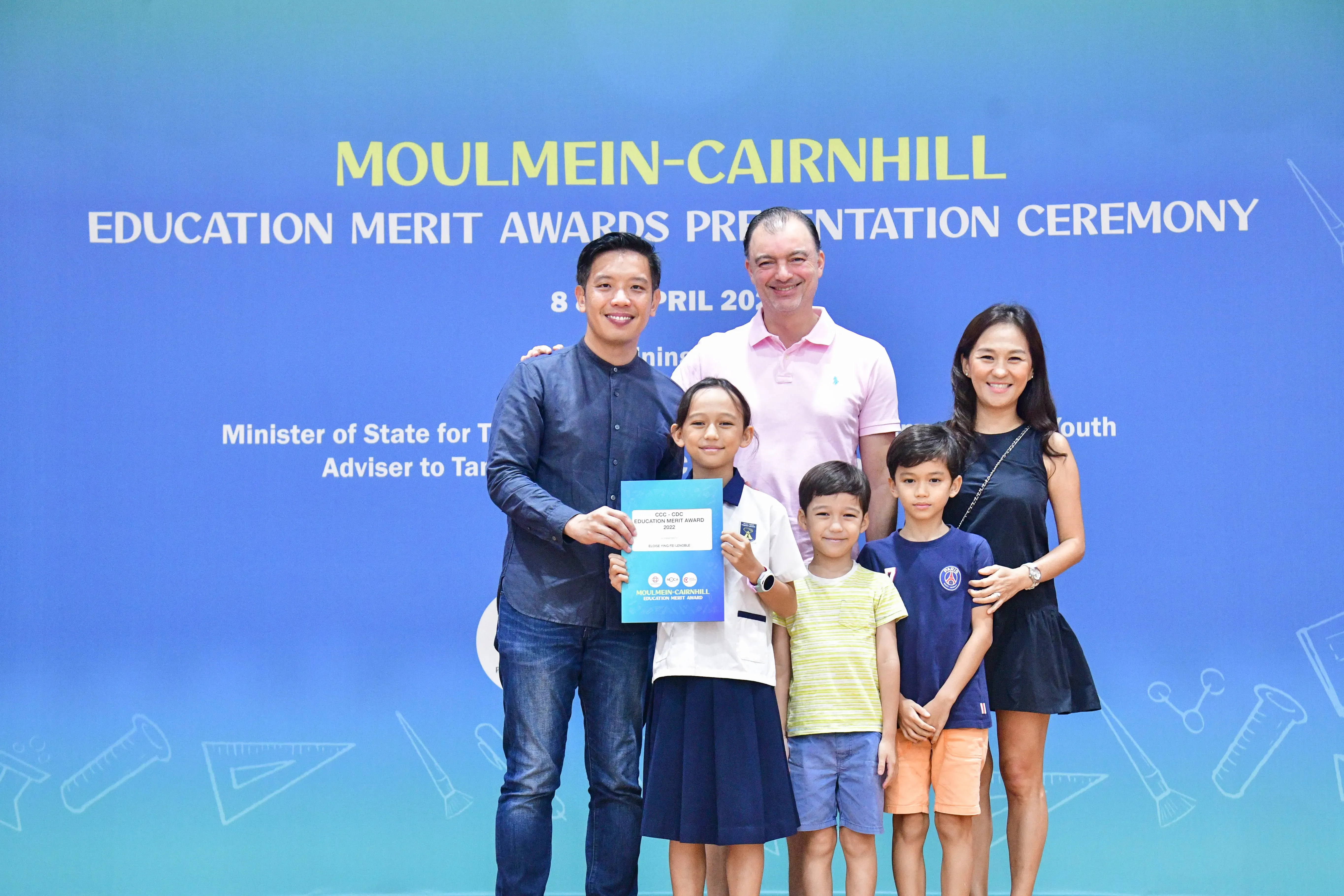 A girl holds an award certificate with three adults and two boys at the Moulmein-Cairnhill Education Merit Awards ceremony.