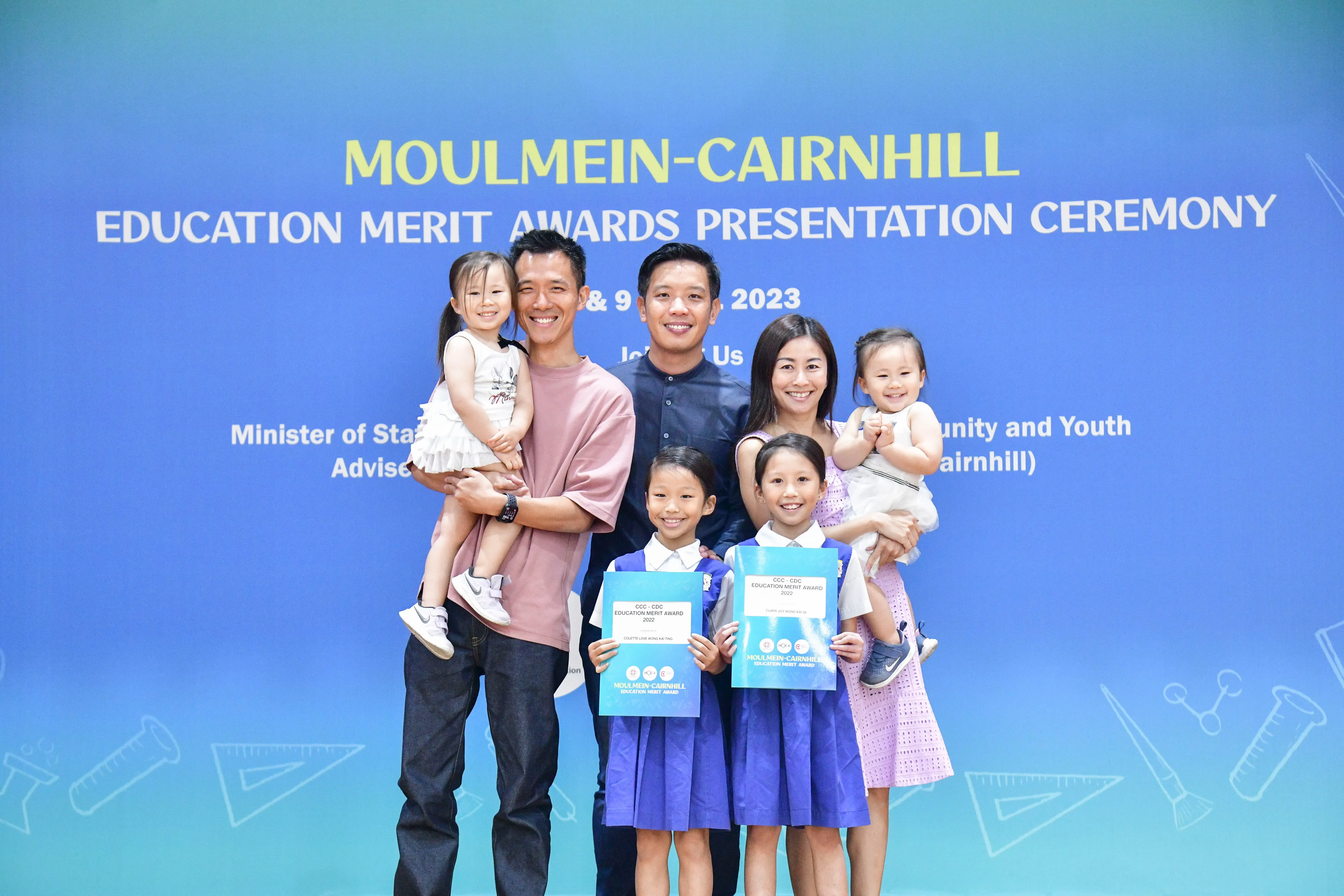 Family of six poses in front of Moulmein-Cairnhill Education Merit Awards 2023 banner, two girls holding award certificates.
