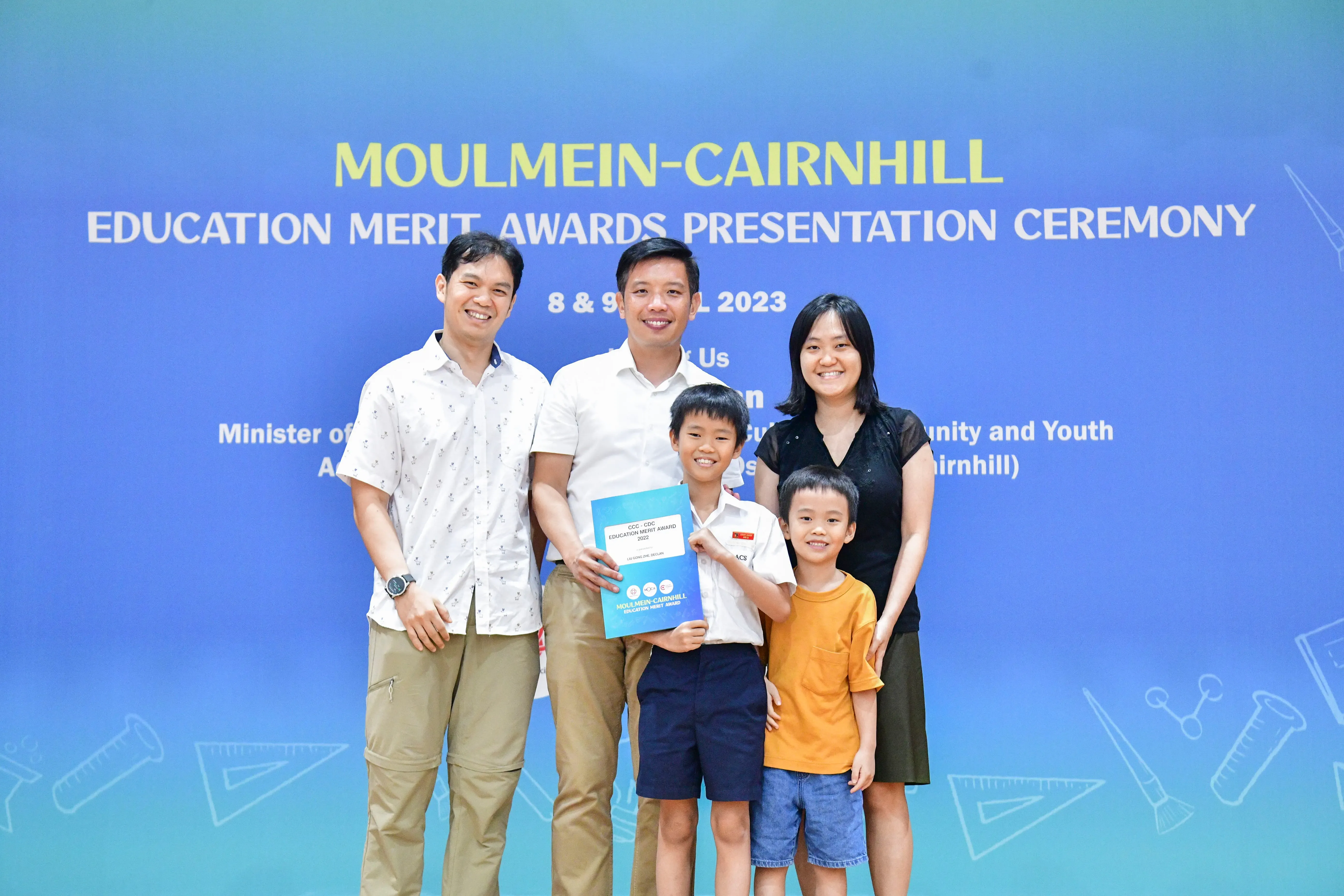 Three adults and two children pose in front of a Moulmein-Cairnhill Education Merit Awards 2023 banner.