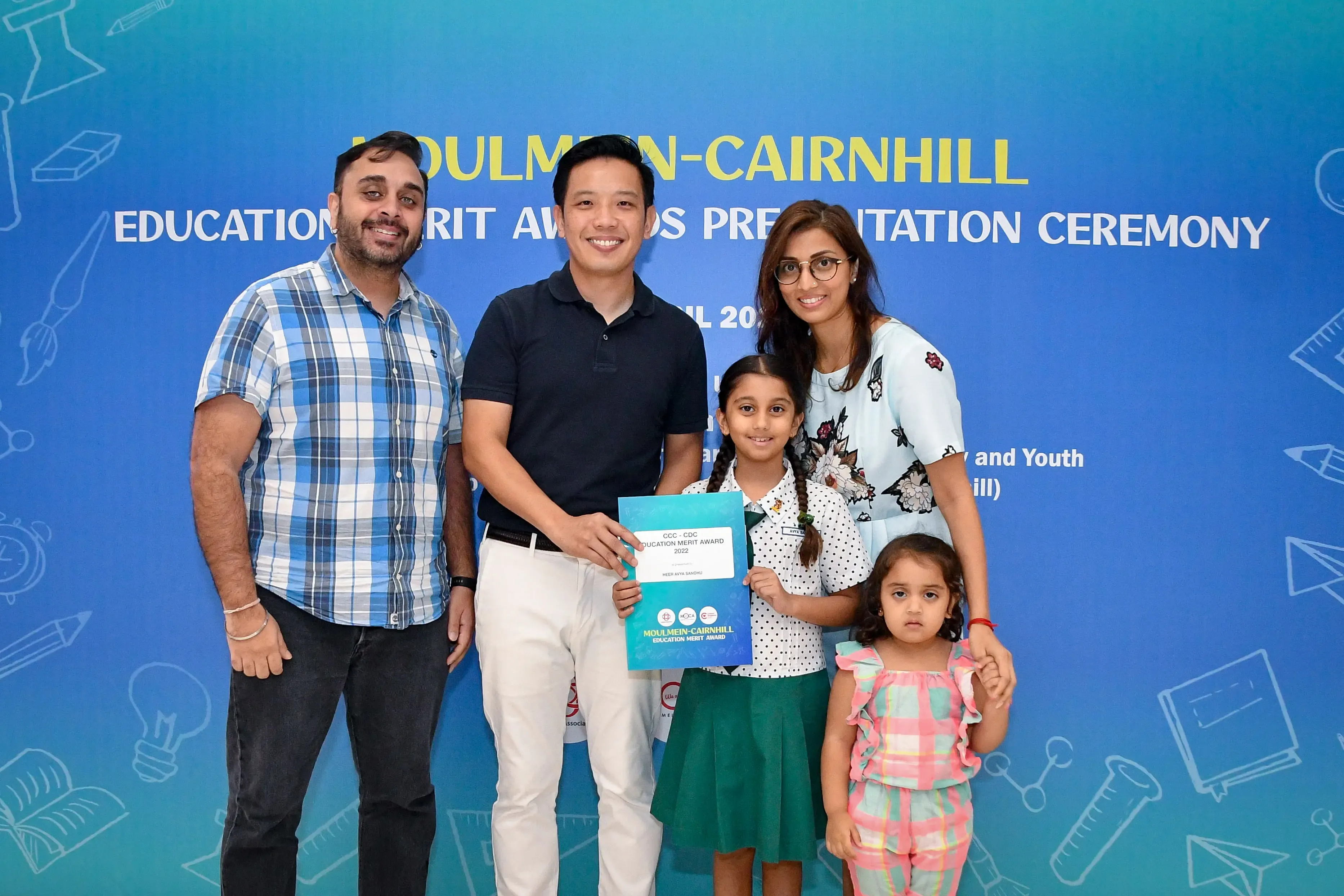 Five people pose at the Moulmein-Cairnhill Education Merit Awards ceremony, with a girl holding a certificate.