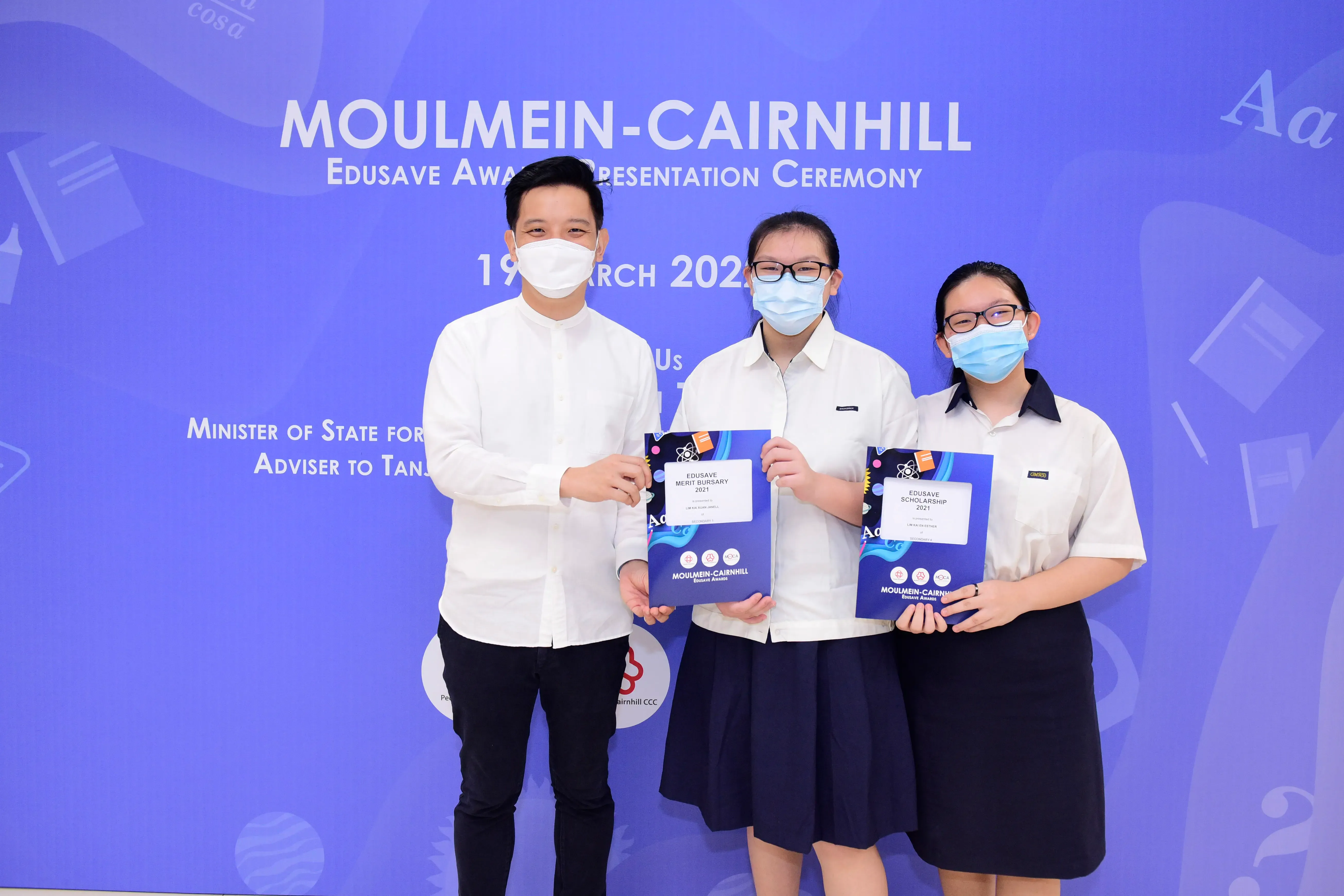 Three masked people pose at the Moulmein-Cairnhill Edusave Award Ceremony, two students holding award certificates.