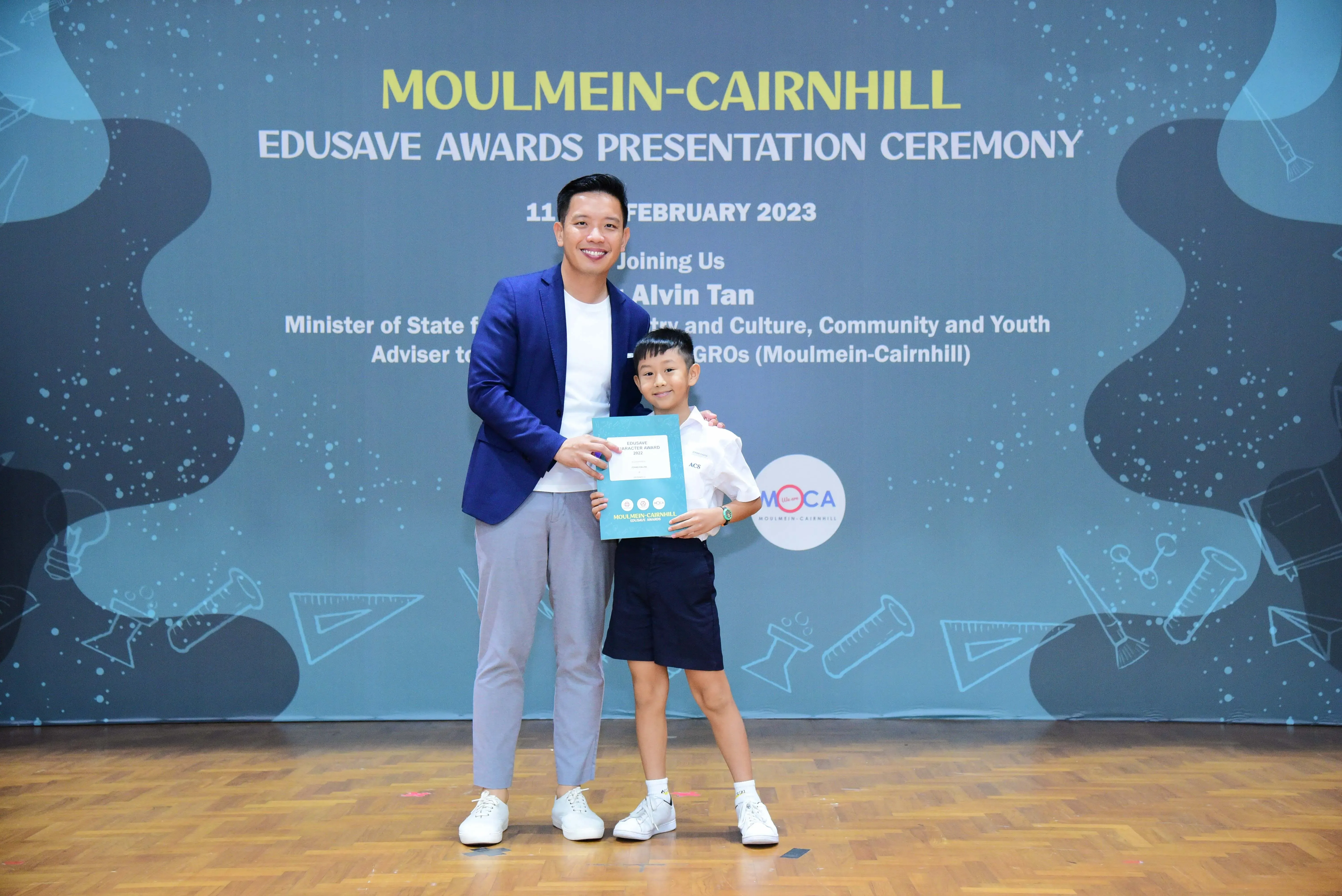 A man in a blue blazer and a young boy hold a certificate at the Moulmein-Cairnhill Edusave Awards Ceremony 2023.