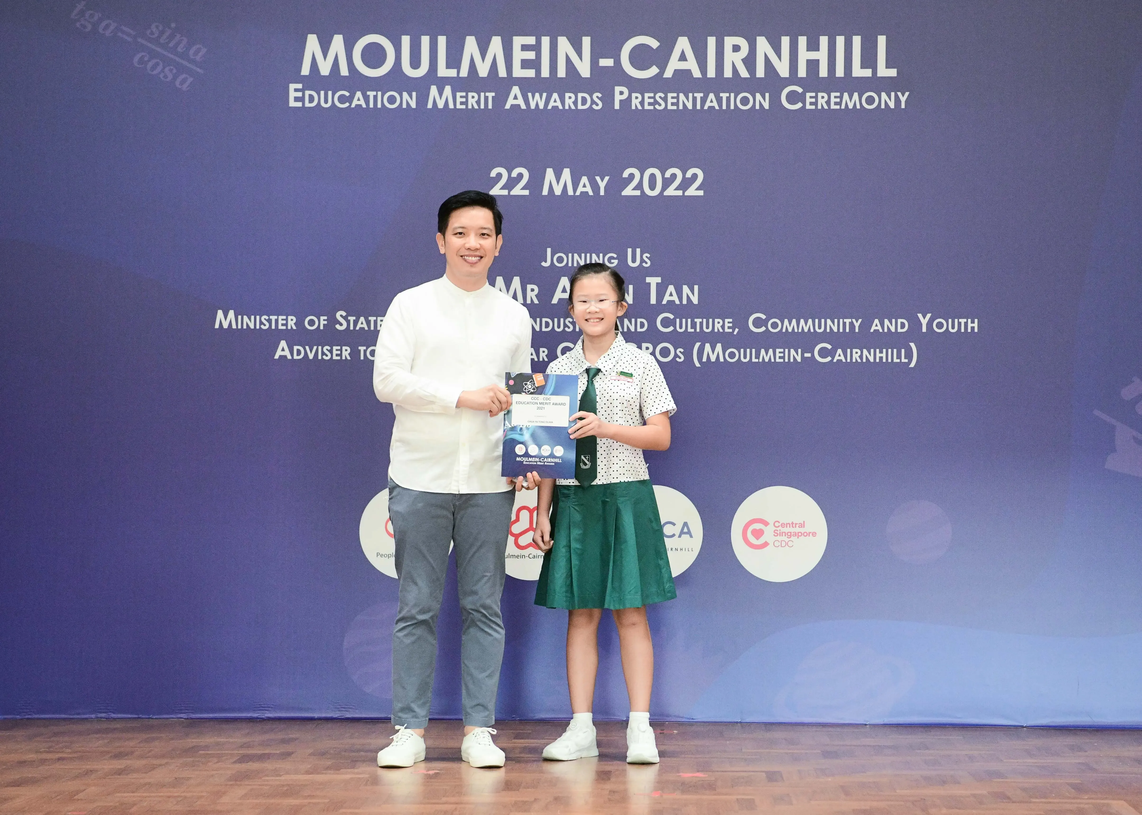 A man and a young girl in school uniform pose together holding an award at the Moulmein-Cairnhill Education Merit Awards ceremony.