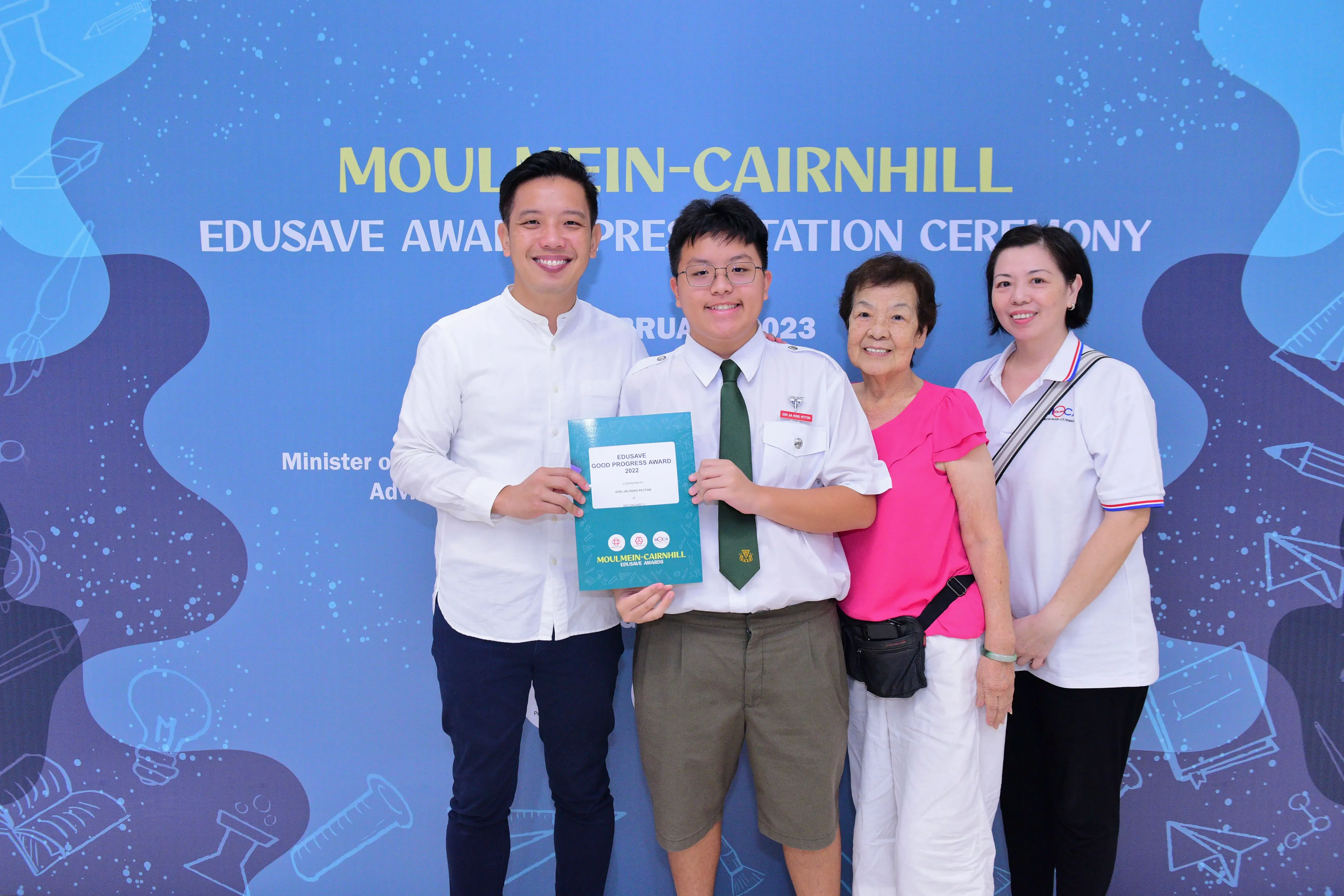 Four people smile at the Moulmein-Cairnhill Edusave Award Presentation Ceremony 2023, with a student holding a certificate.