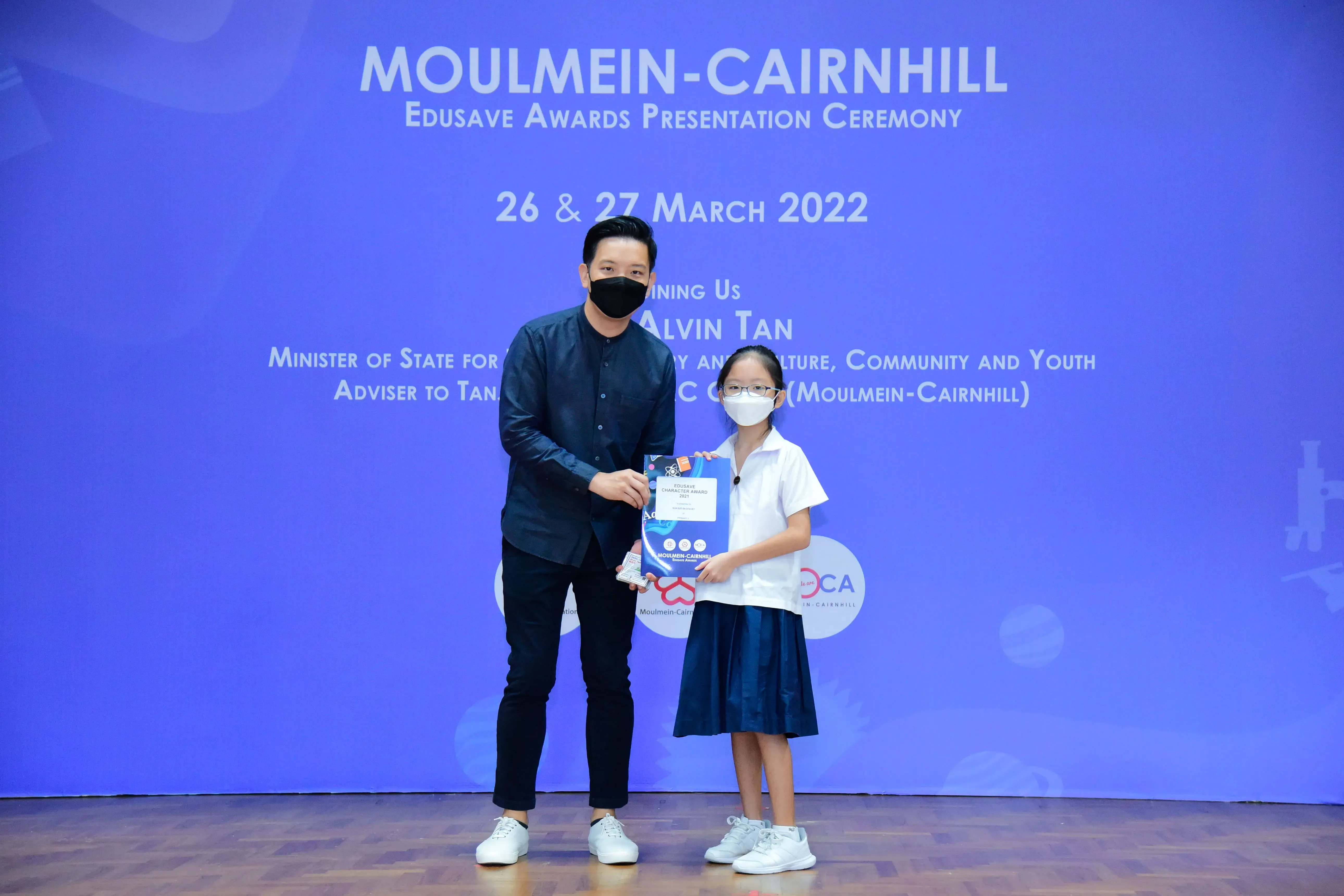 An adult and a young girl in school uniform hold an award folder at the Moulmein-Cairnhill Edusave Awards Ceremony 2022.