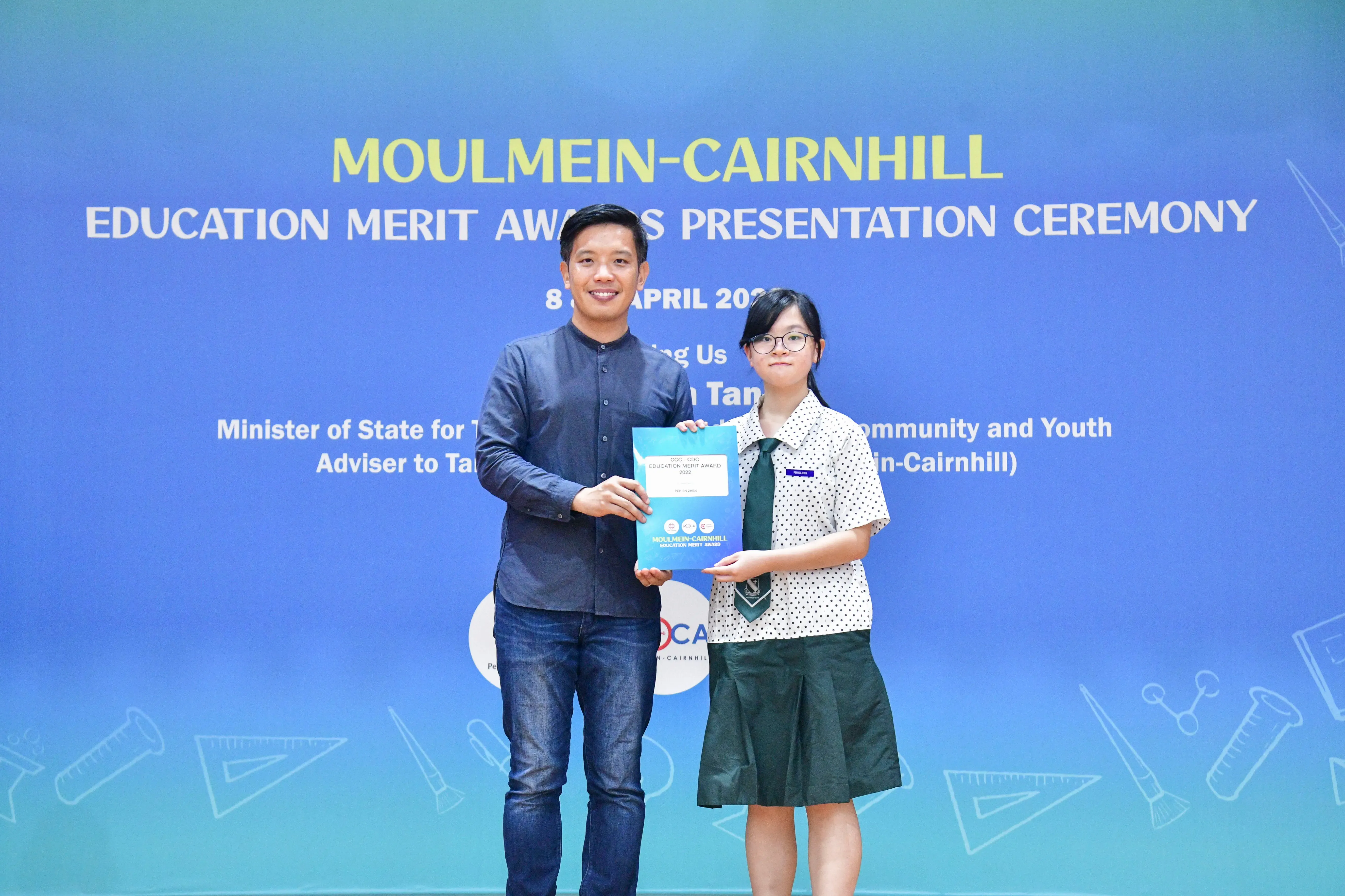 A man and a young girl in school uniform hold a certificate together at the Moulmein-Cairnhill Education Merit Awards ceremony.