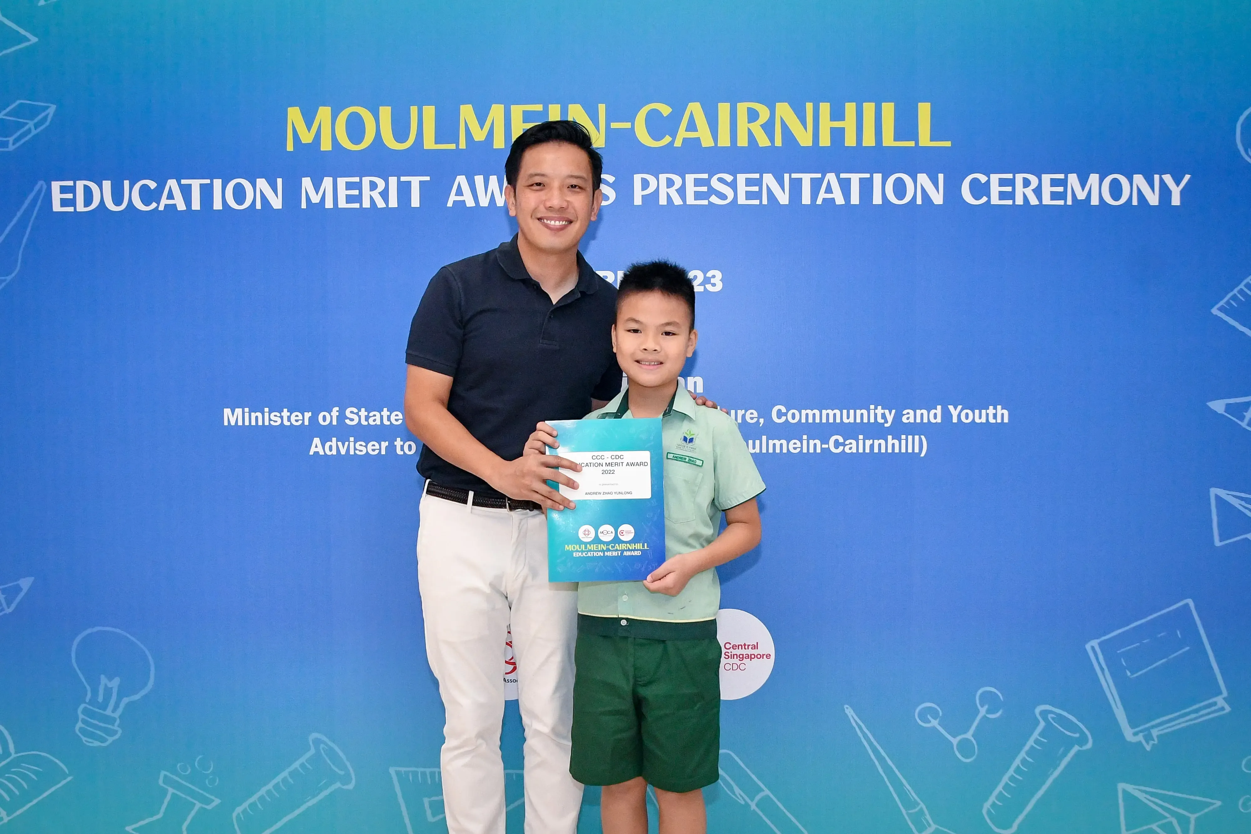 A man and a young boy holding a merit award certificate at the Moulmein-Cairnhill Education Merit Awards Ceremony.
