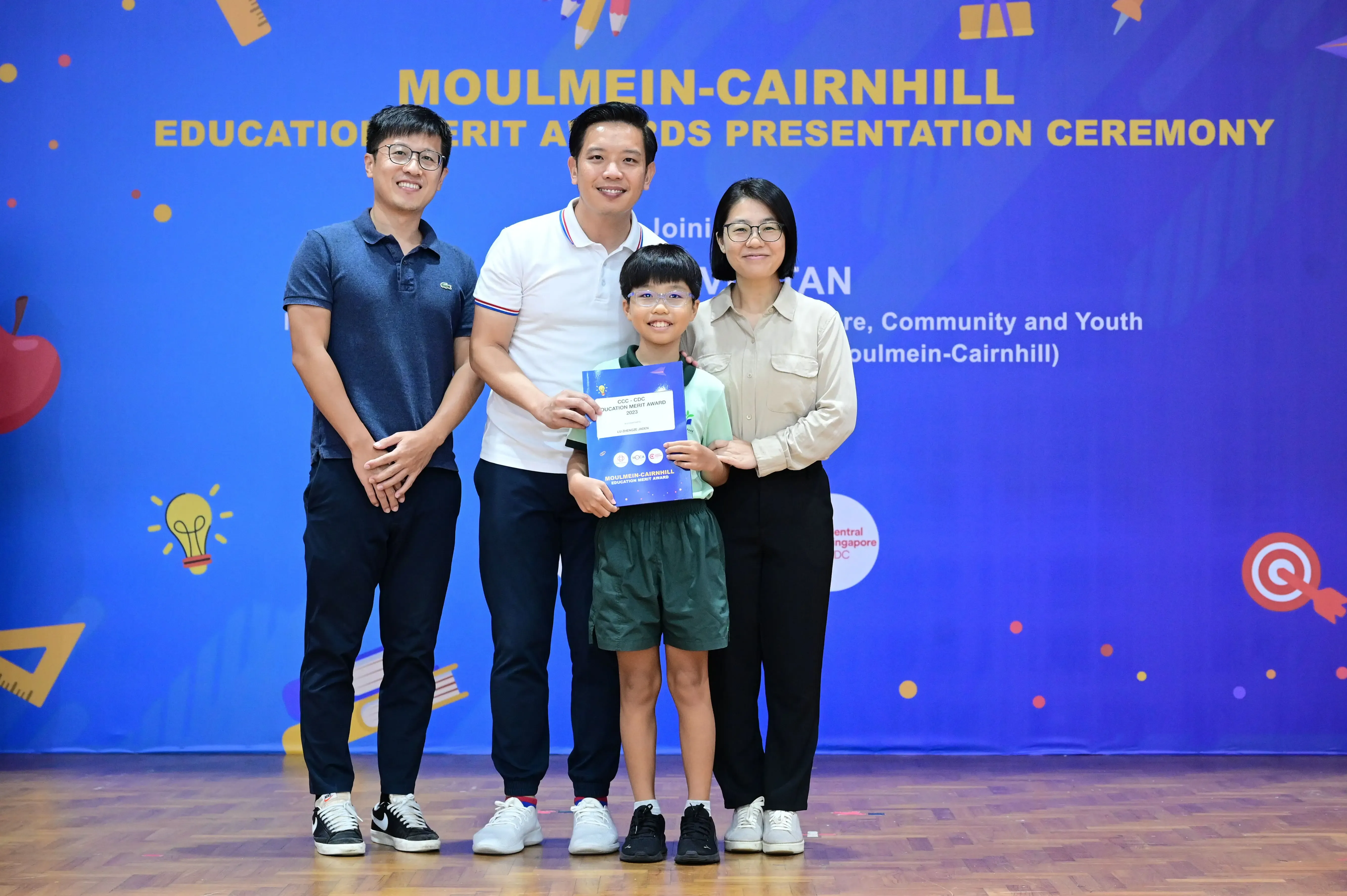Three adults and a child pose at the Moulmein-Cairnhill Education Merit Awards ceremony, child holding a certificate.