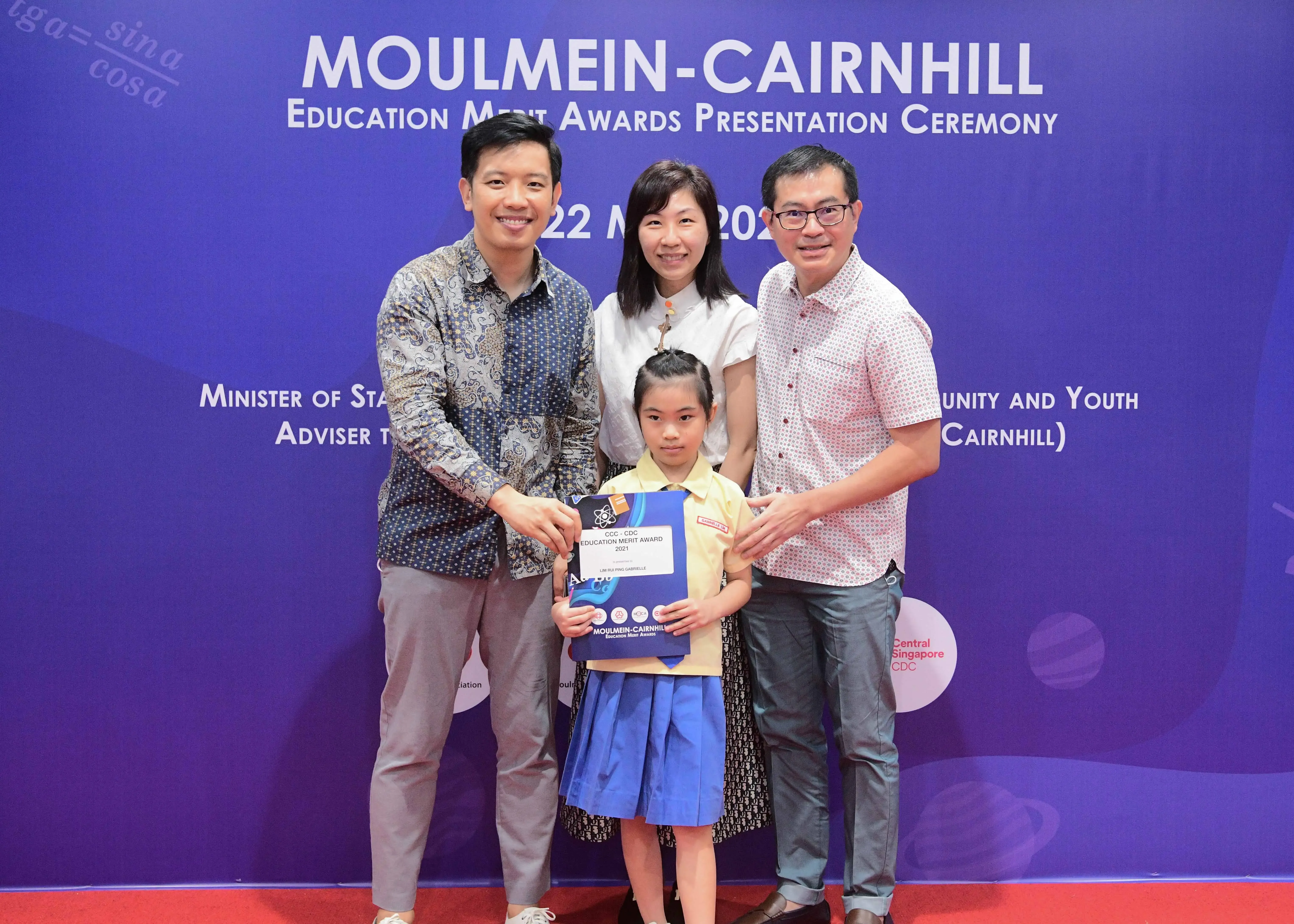 Three adults and a young girl holding an award certificate at the Moulmein-Cairnhill Education Merit Awards ceremony.