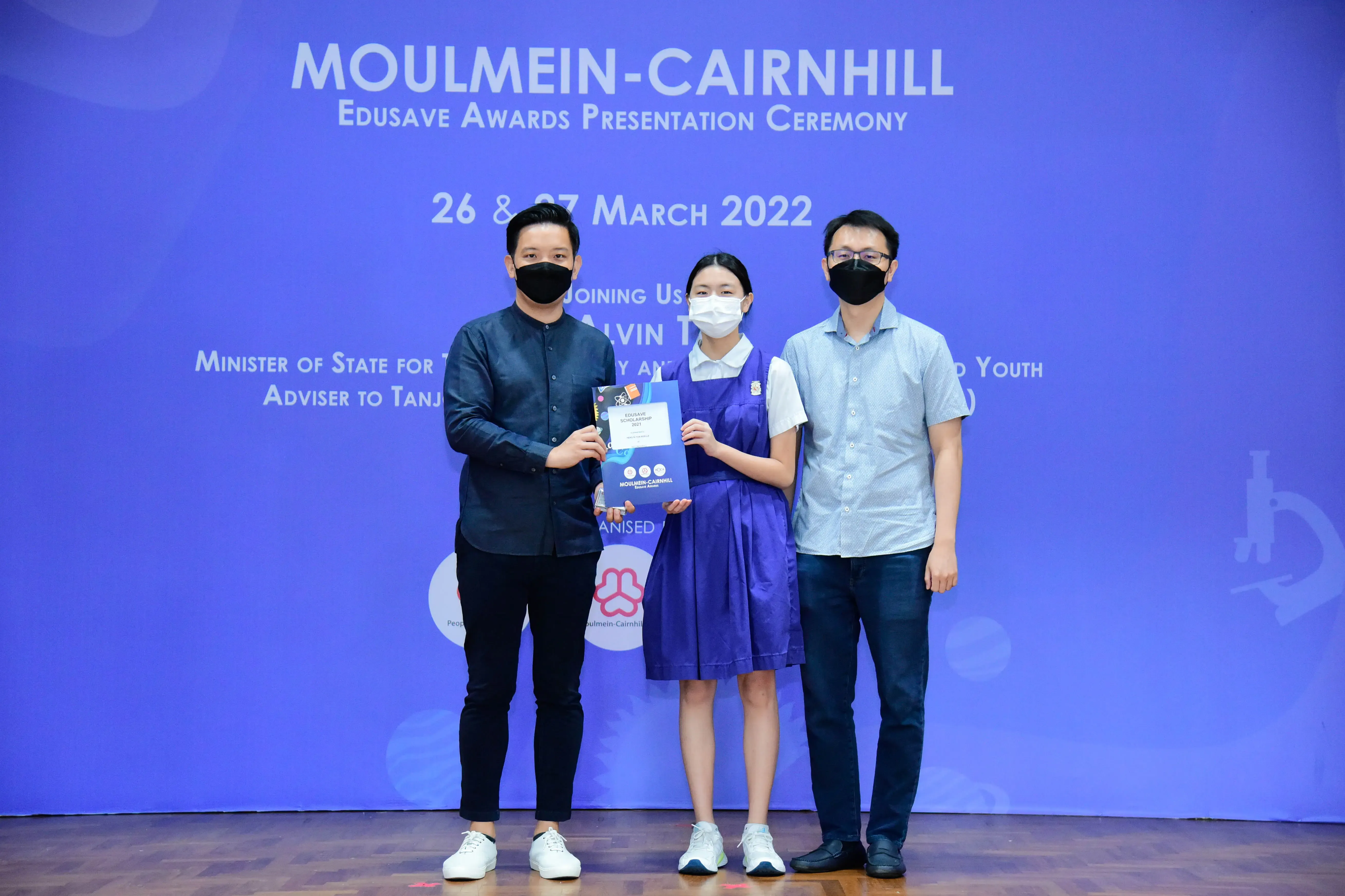 Three masked people pose on a stage at the Moulmein-Cairnhill Edusave Awards Presentation Ceremony, March 2022.