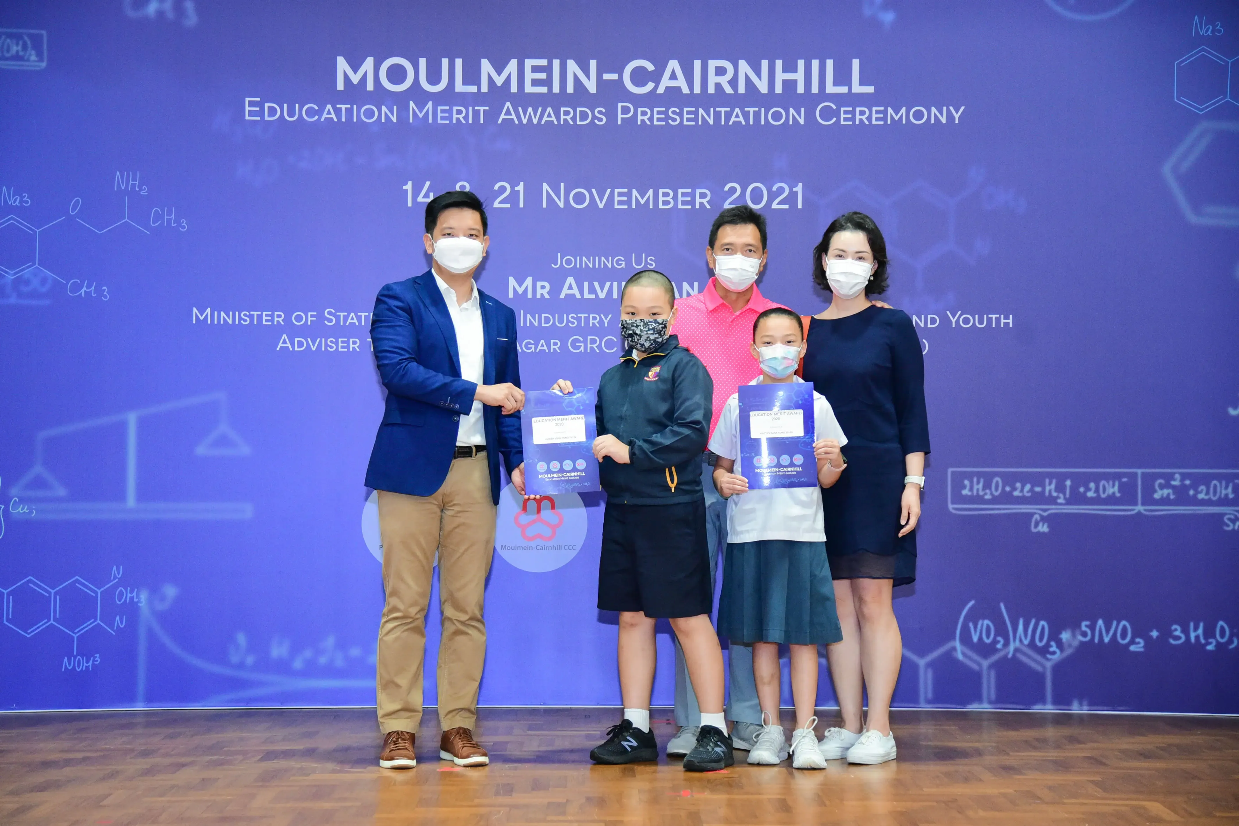 Five masked people pose at the Moulmein-Cairnhill Education Merit Awards Presentation Ceremony, two children holding awards.
