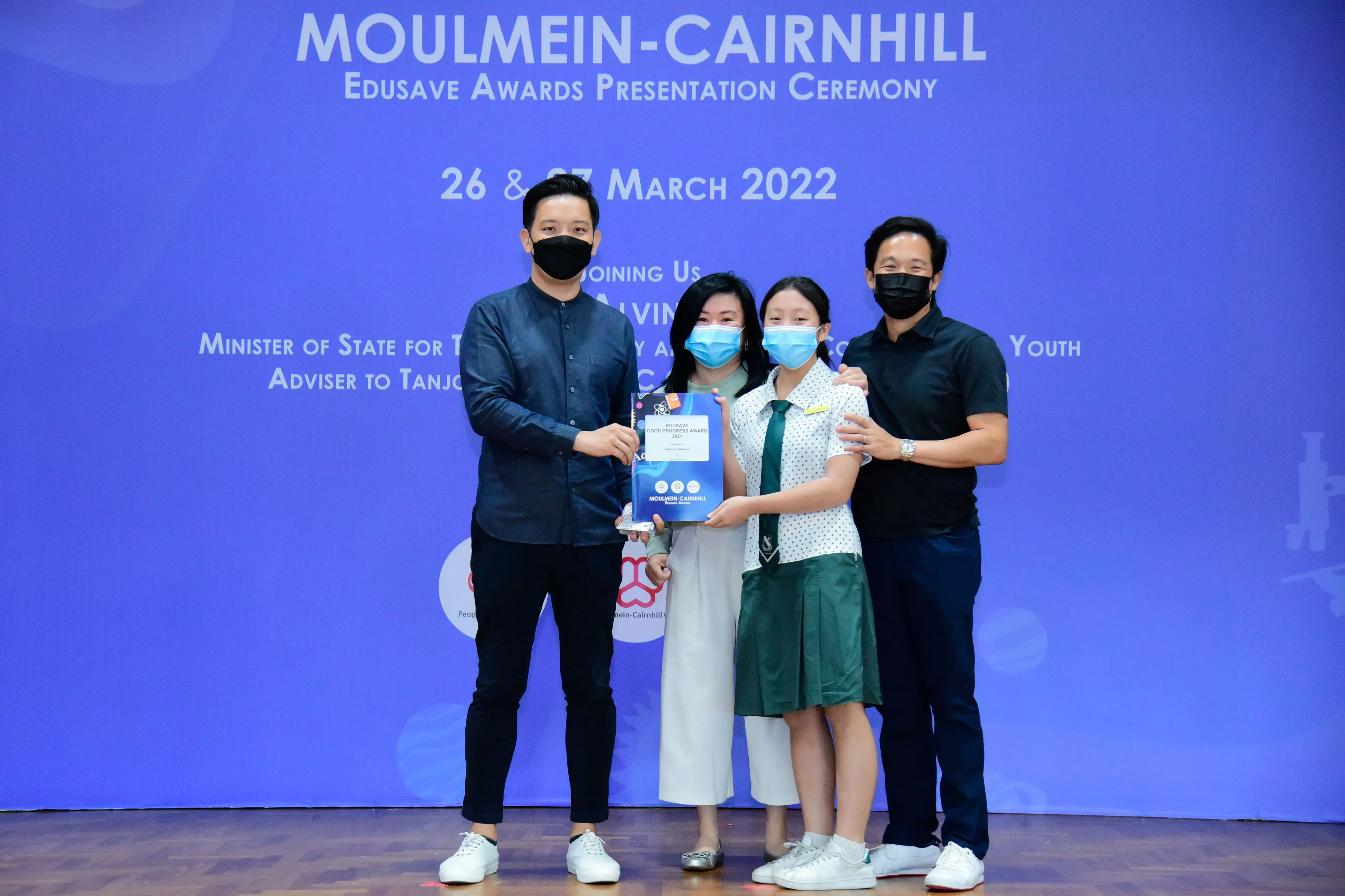Four masked people pose on stage at the Moulmein-Cairnhill Edusave Awards Presentation Ceremony, holding an award.