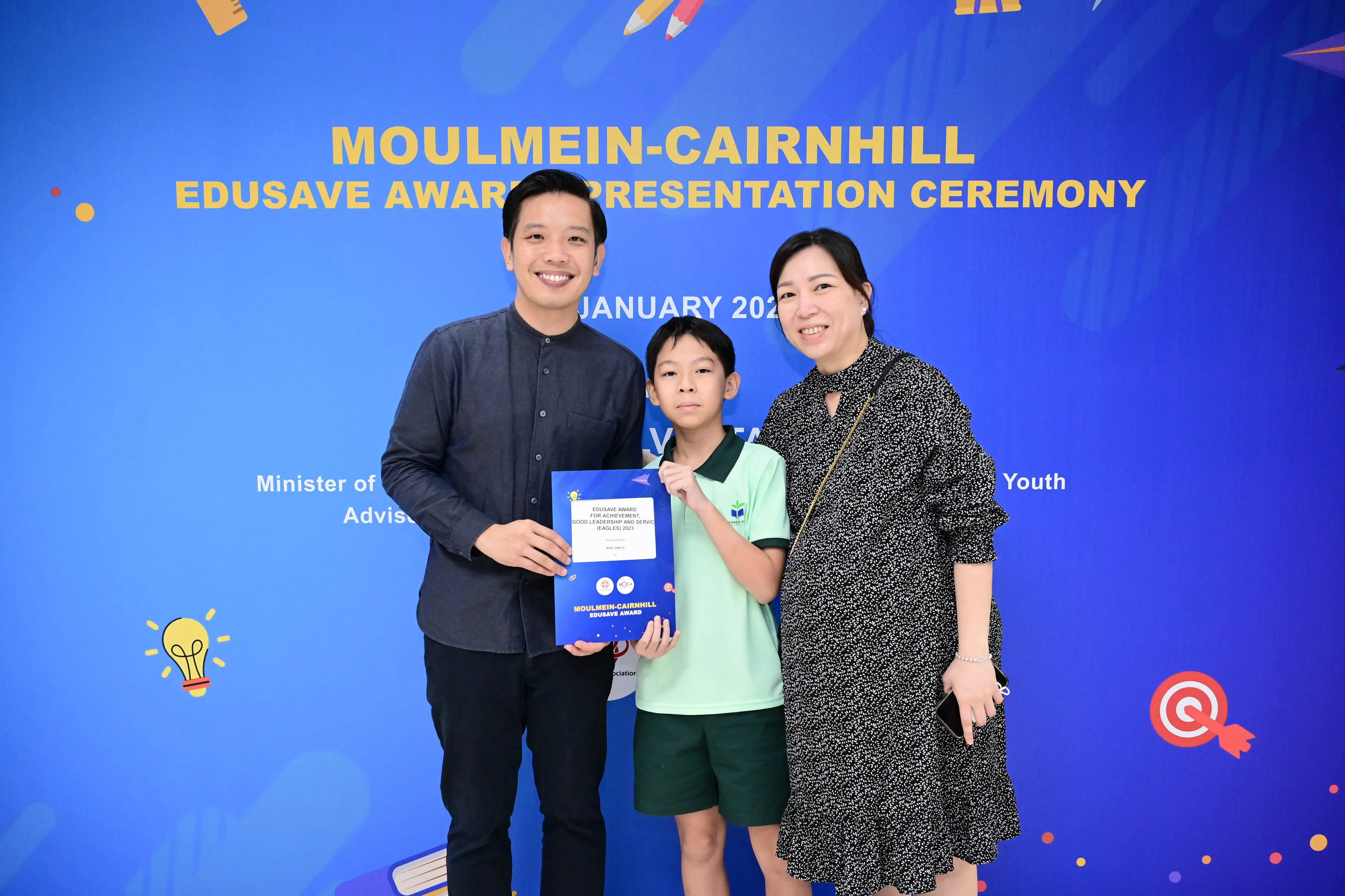 A boy holding an award certificate stands between two smiling adults at the Moulmein-Cairnhill Edusave Award Presentation Ceremony.