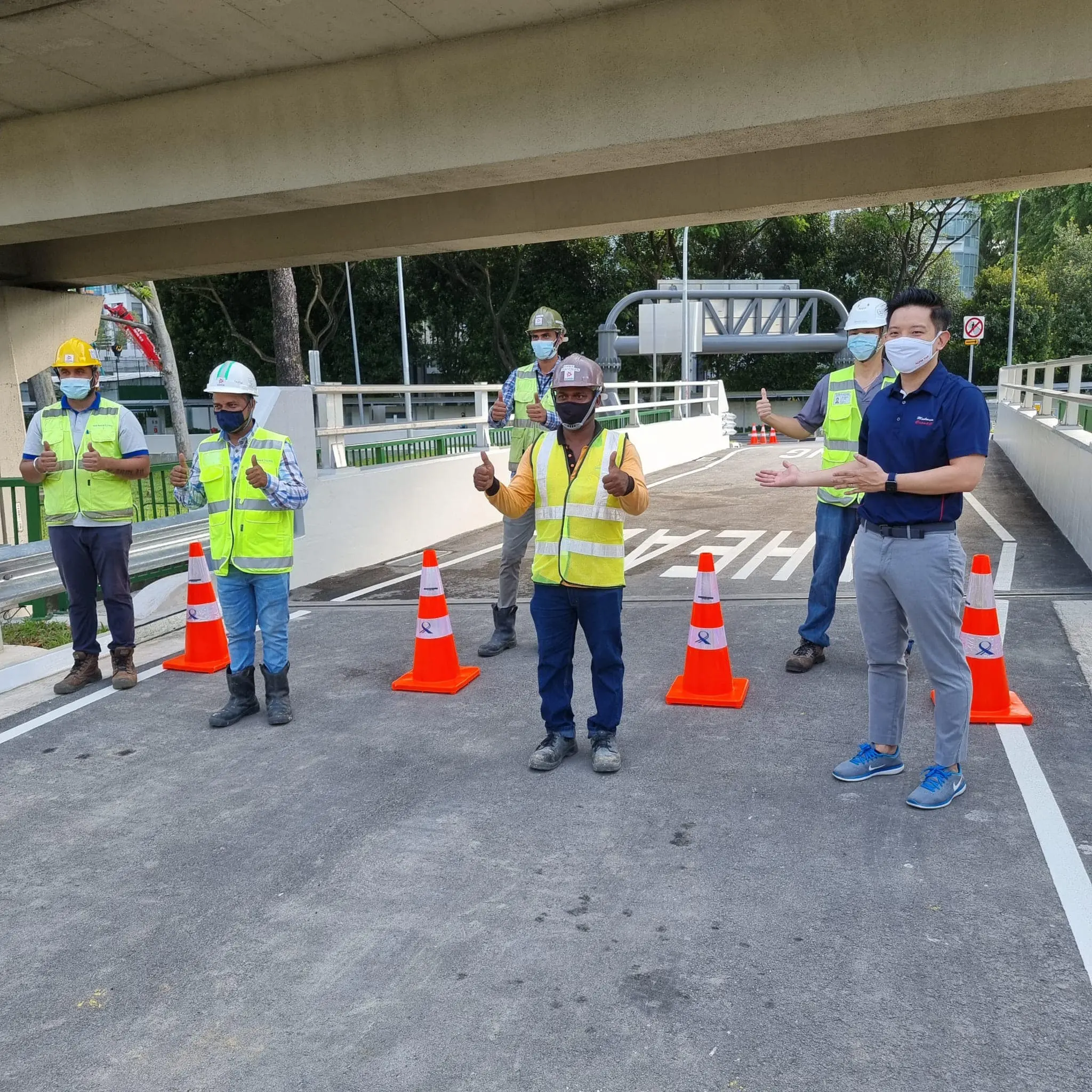 Six masked workers in safety vests stand on a road under a bridge, surrounded by orange traffic cones, giving thumbs up.