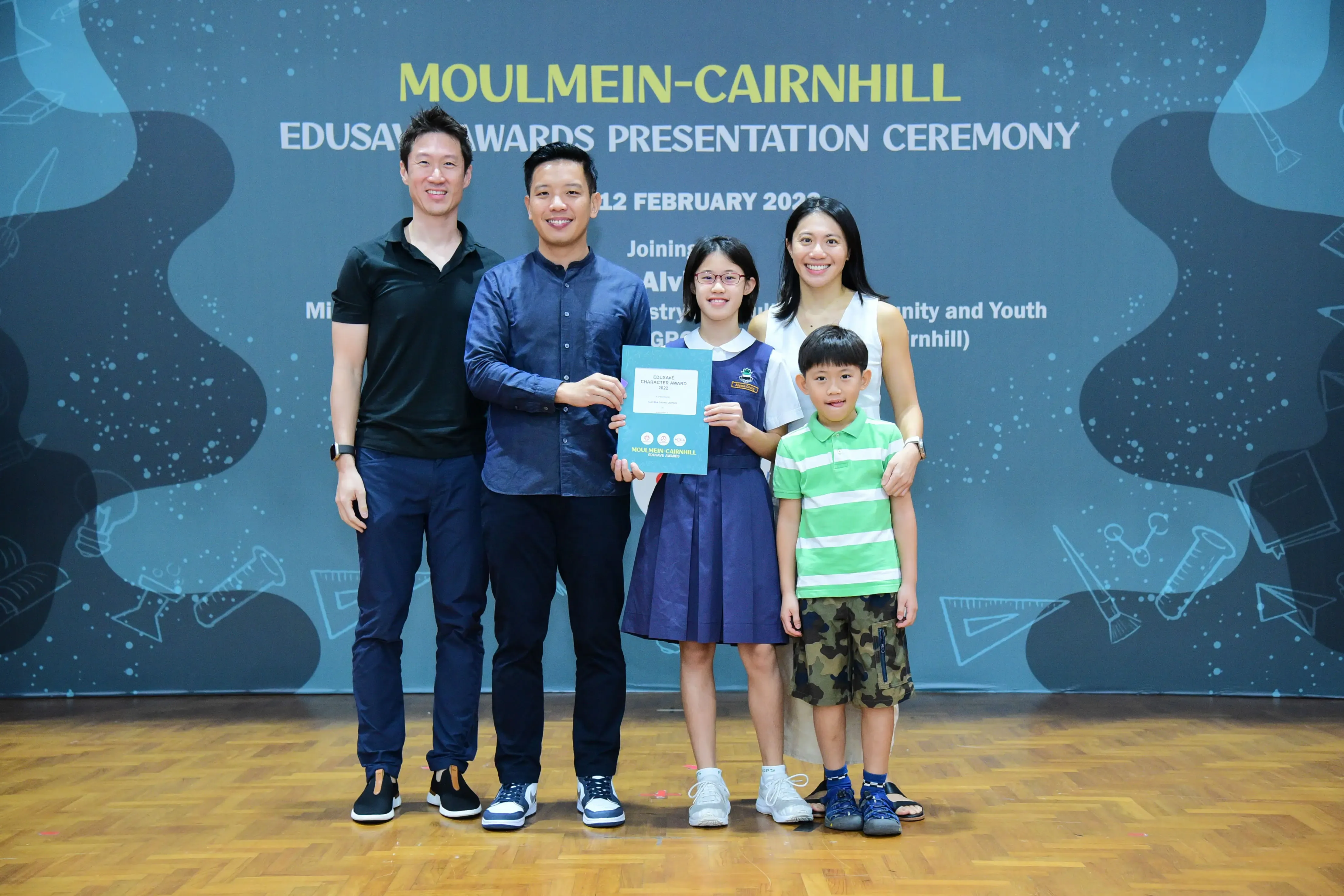 Five people pose at the Moulmein-Cairnhill EduSave Awards Presentation Ceremony, with a girl holding a certificate.