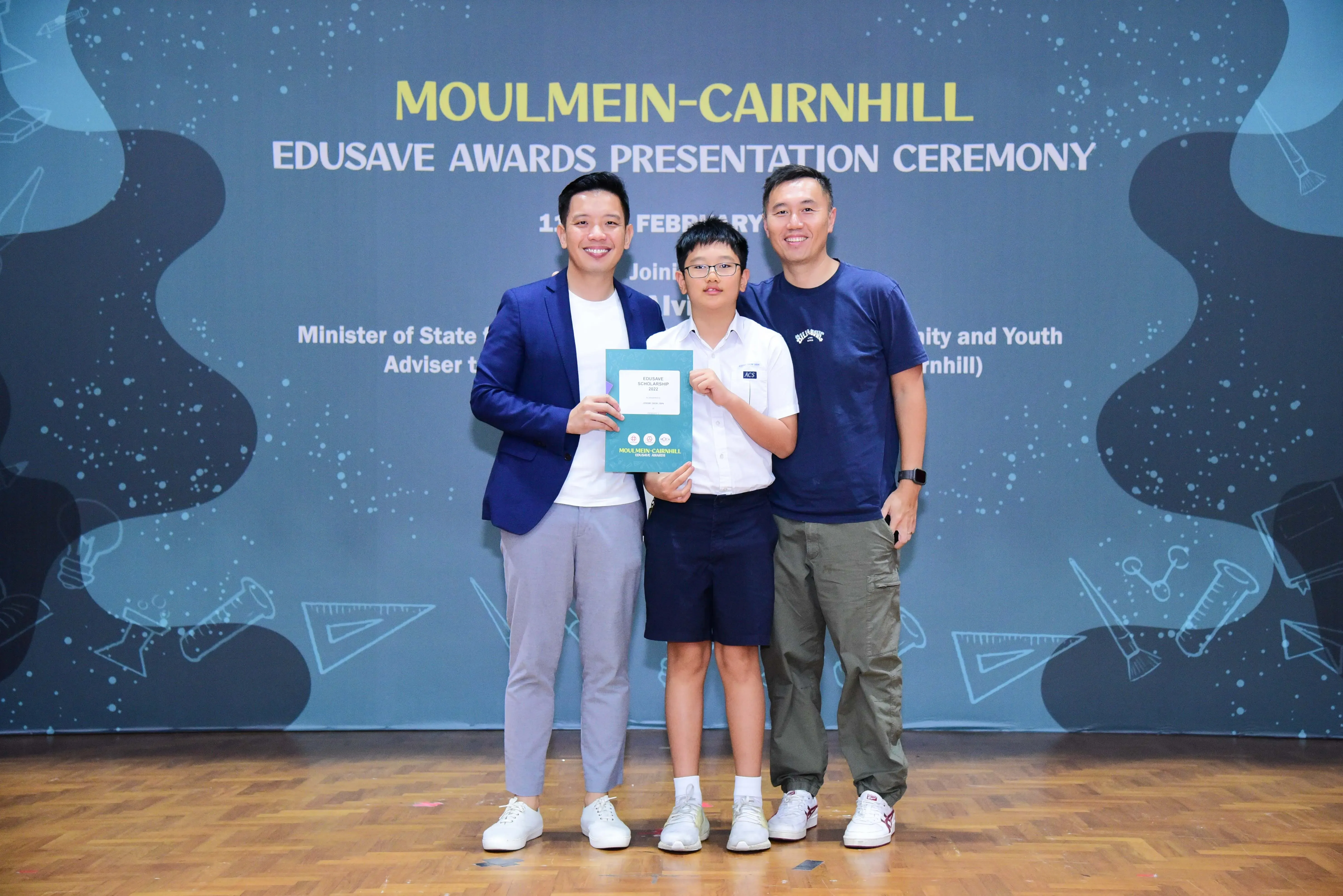 Three people pose smiling at the Moulmein-Cairnhill Edusave Awards Presentation Ceremony, a boy holding a certificate.