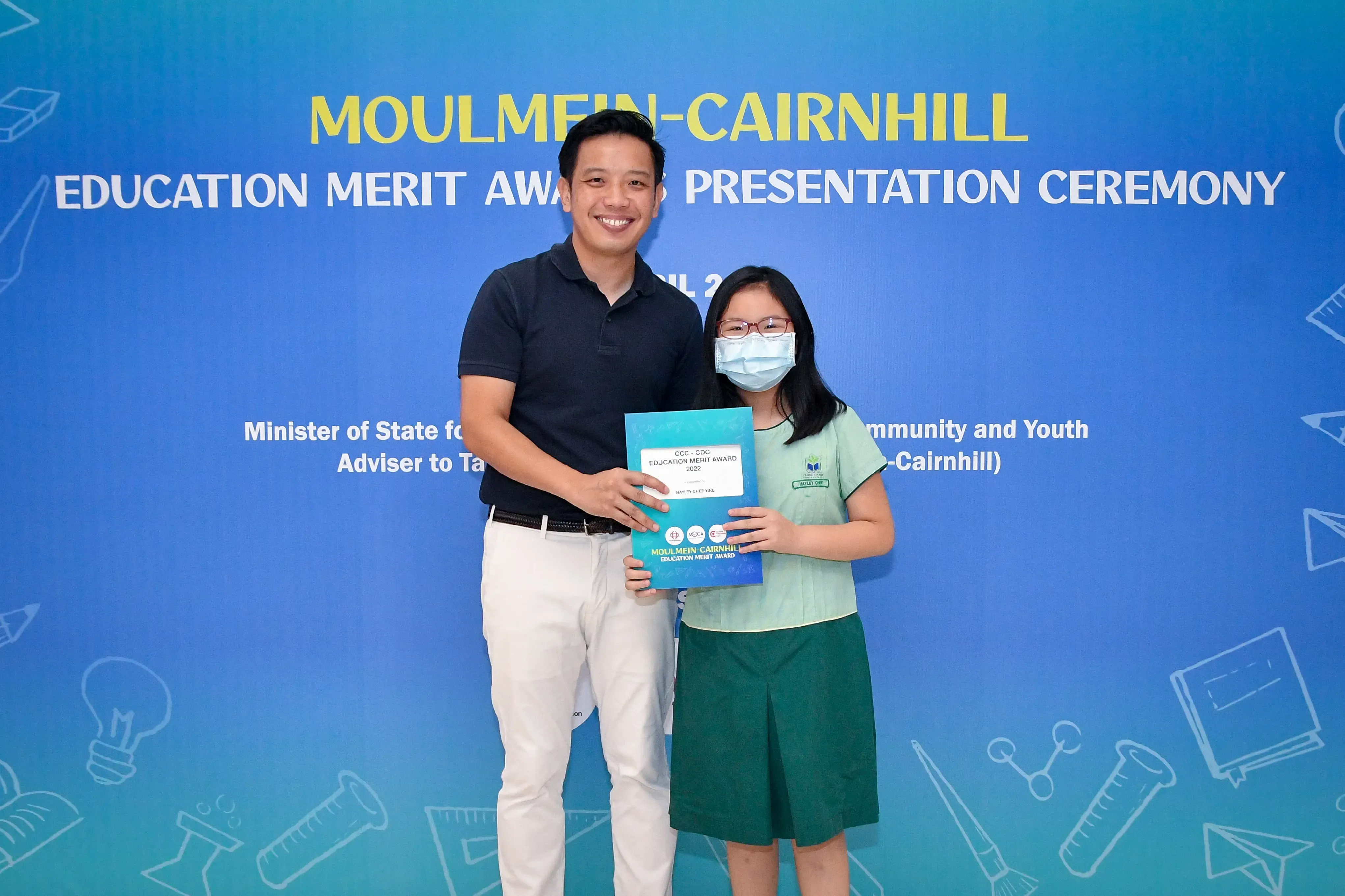 A man and a masked girl hold an award certificate at the Moulmein-Cairnhill Education Merit Award Presentation Ceremony.
