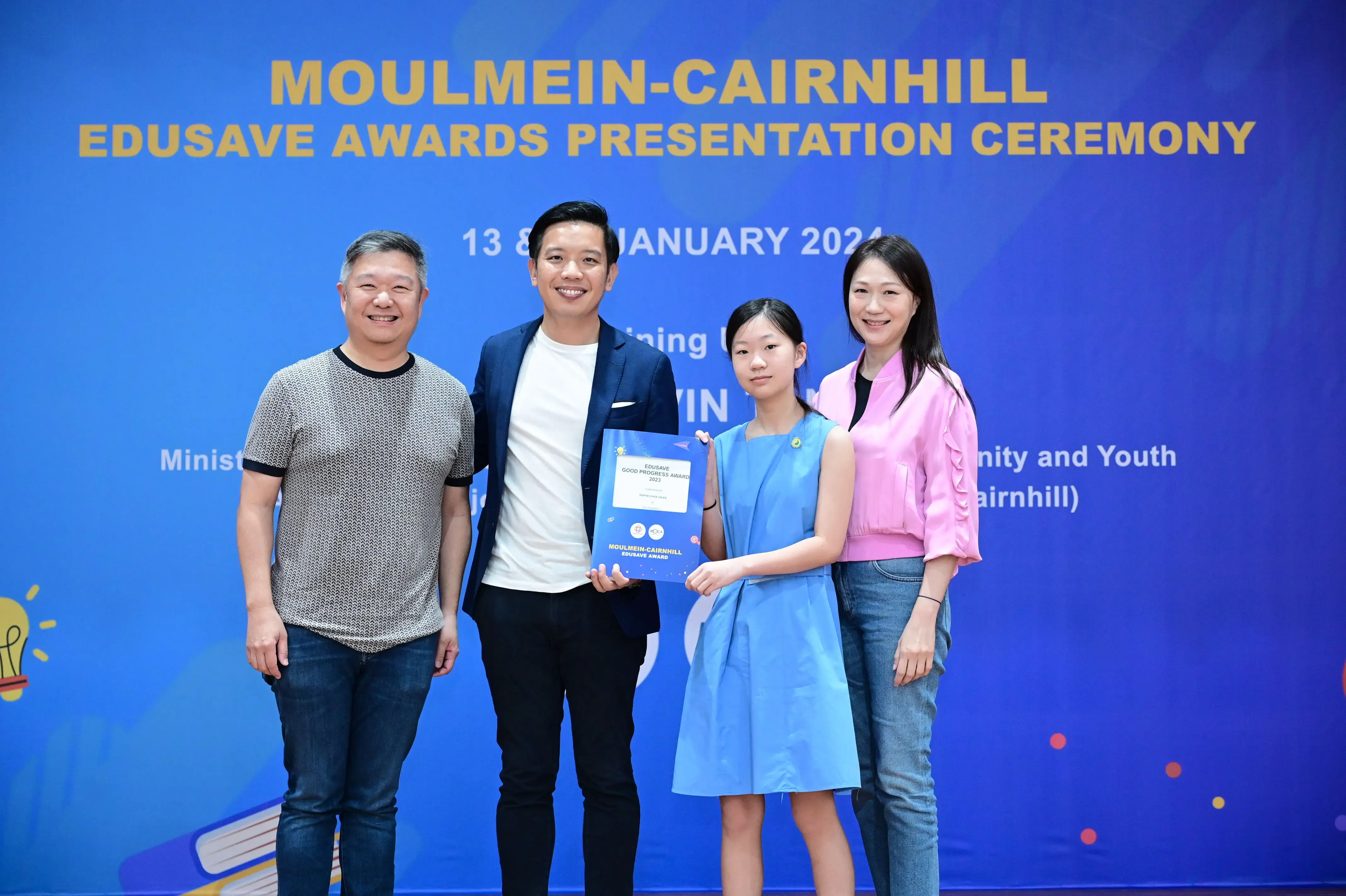 Four people pose in front of a blue Moulmein-Cairnhill Edusave Awards Presentation Ceremony banner, one holding a certificate.