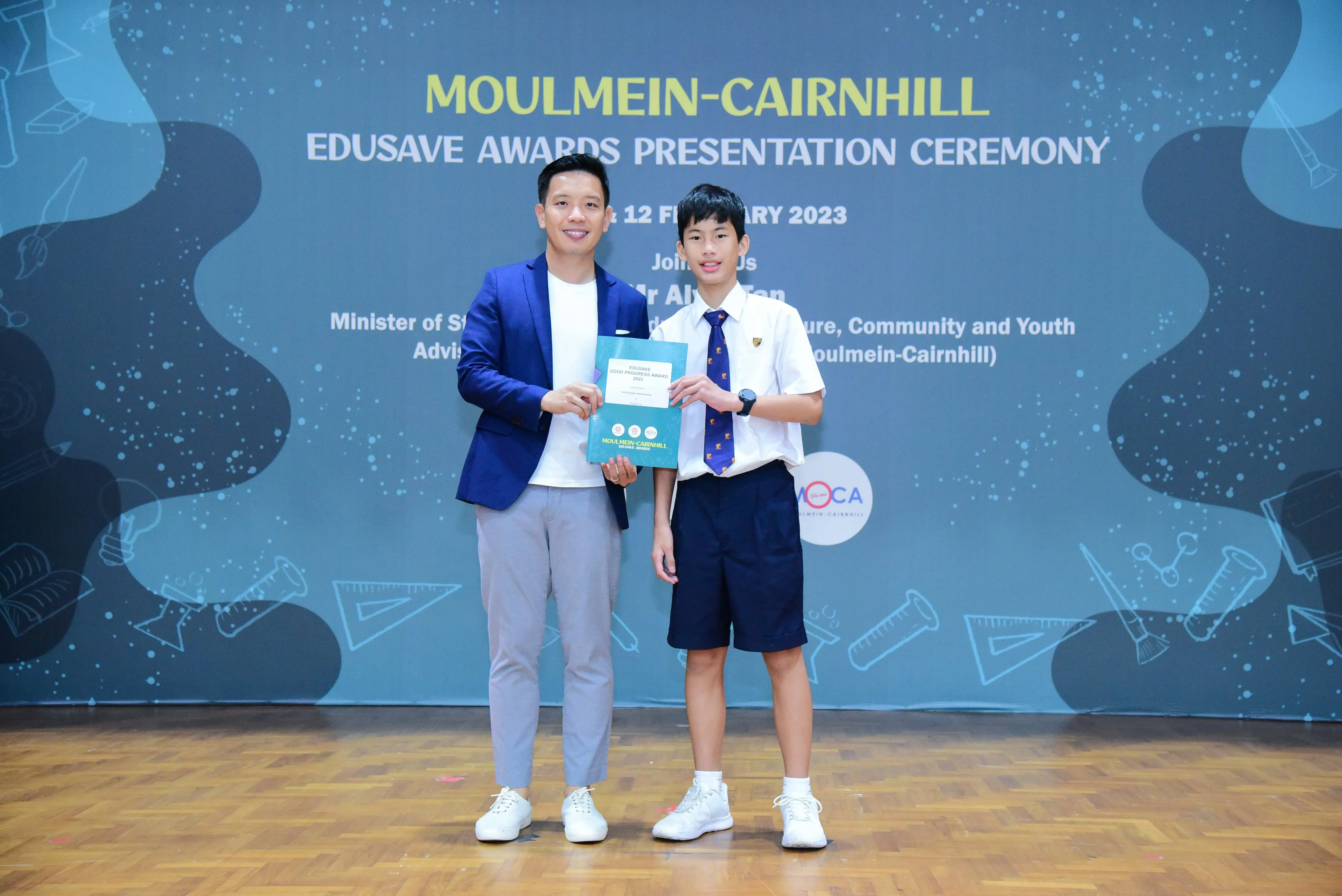An adult in a blue blazer and a young student in uniform hold a certificate at the Moulmein-Cairnhill Edusave Awards Ceremony.