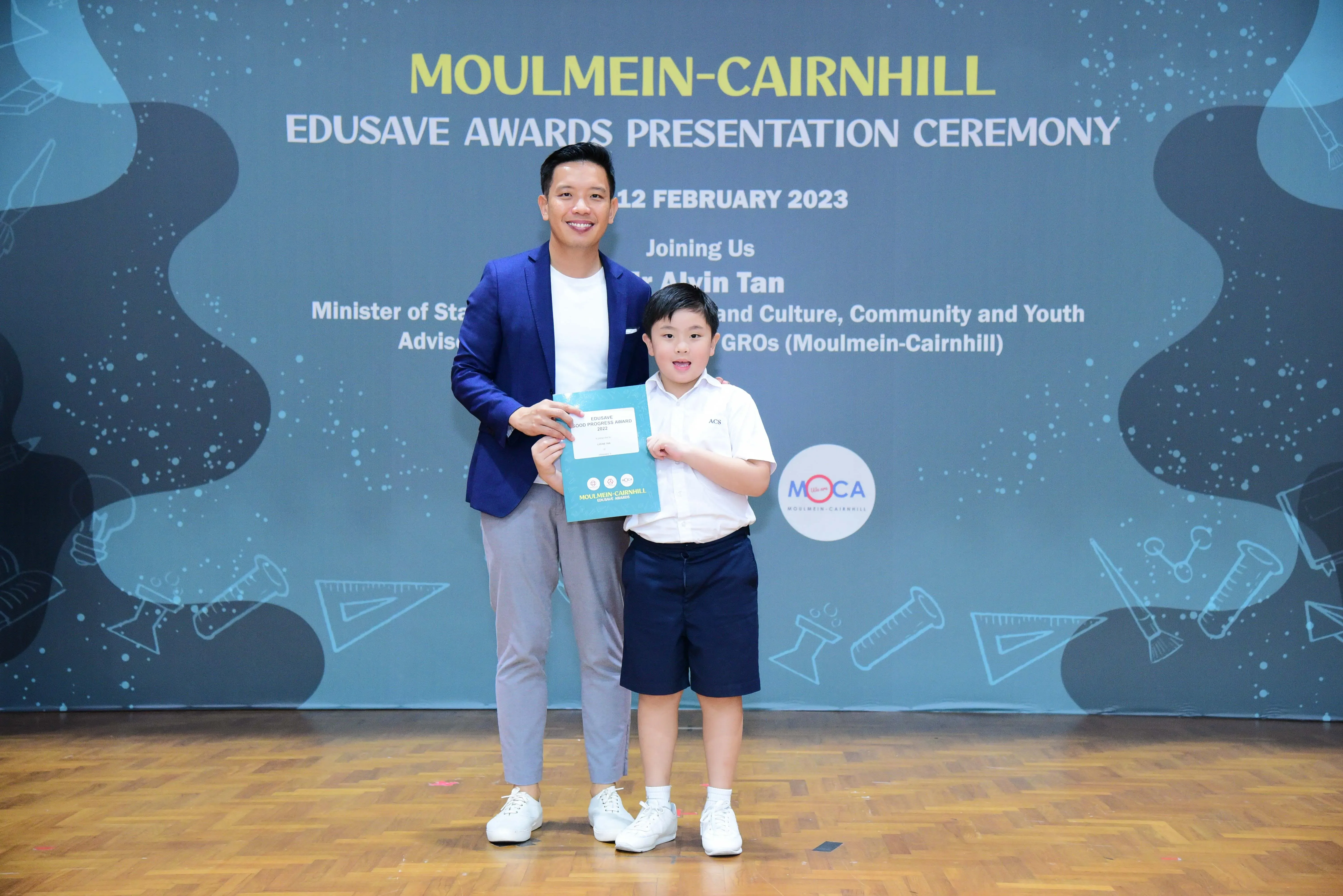 A man in a blue blazer and a young boy in school uniform hold a certificate at the Moulmein-Cairnhill Edusave Awards 2023.