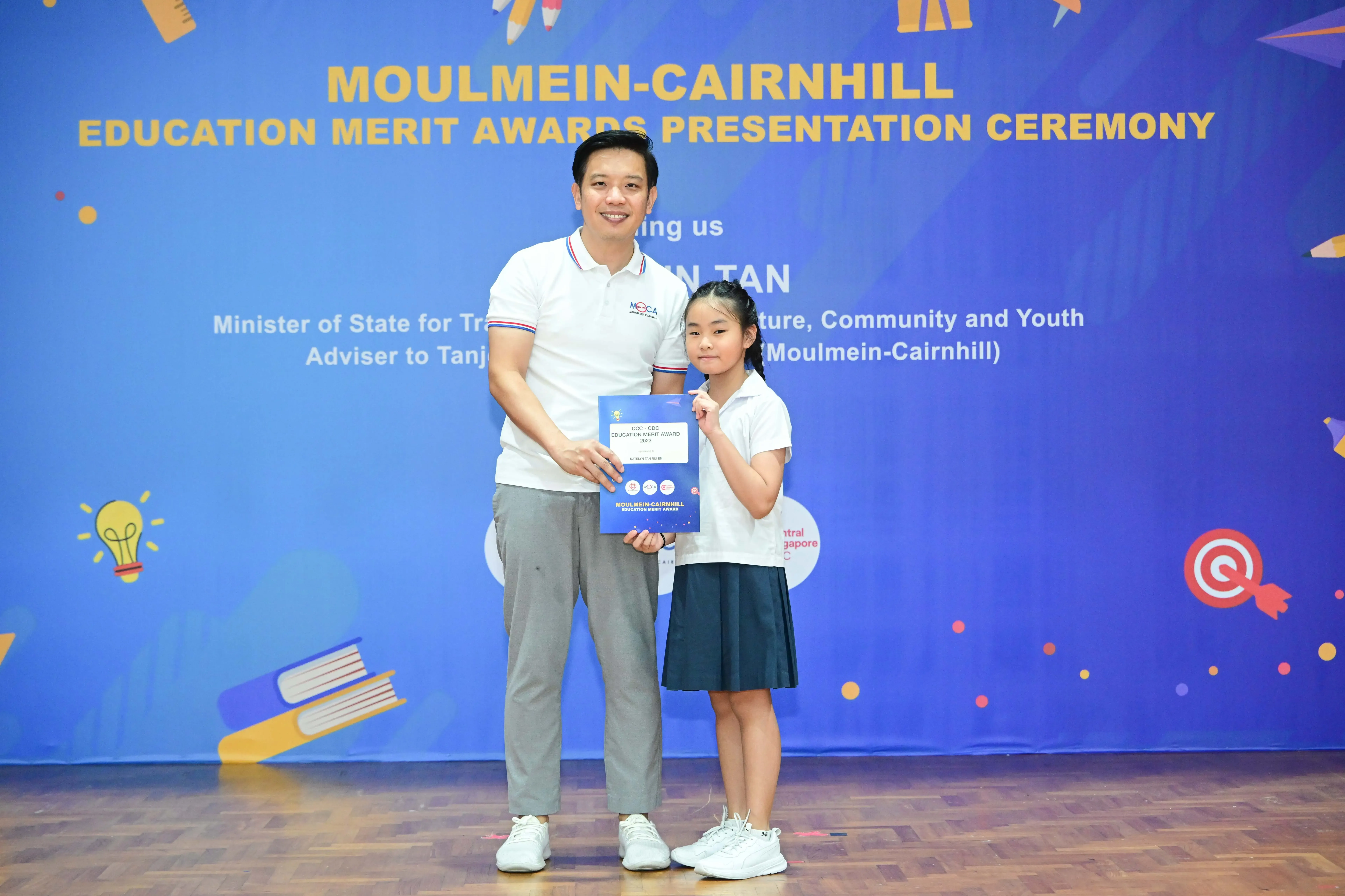 A man and a young girl hold a blue certificate in front of a Moulmein-Cairnhill Education Merit Awards banner.