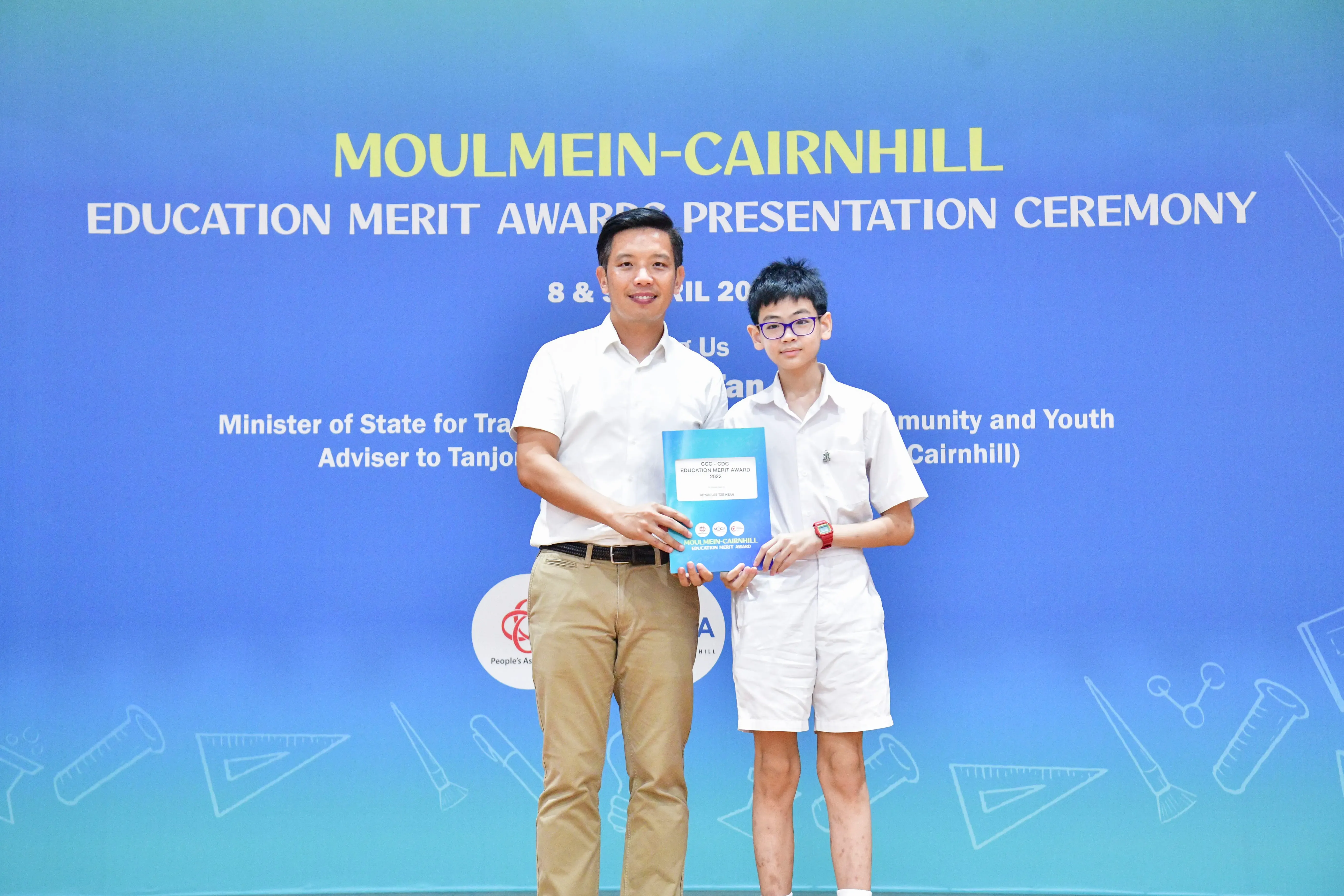 An adult and a young student hold a blue award folder at the Moulmein-Cairnhill Education Merit Awards ceremony.