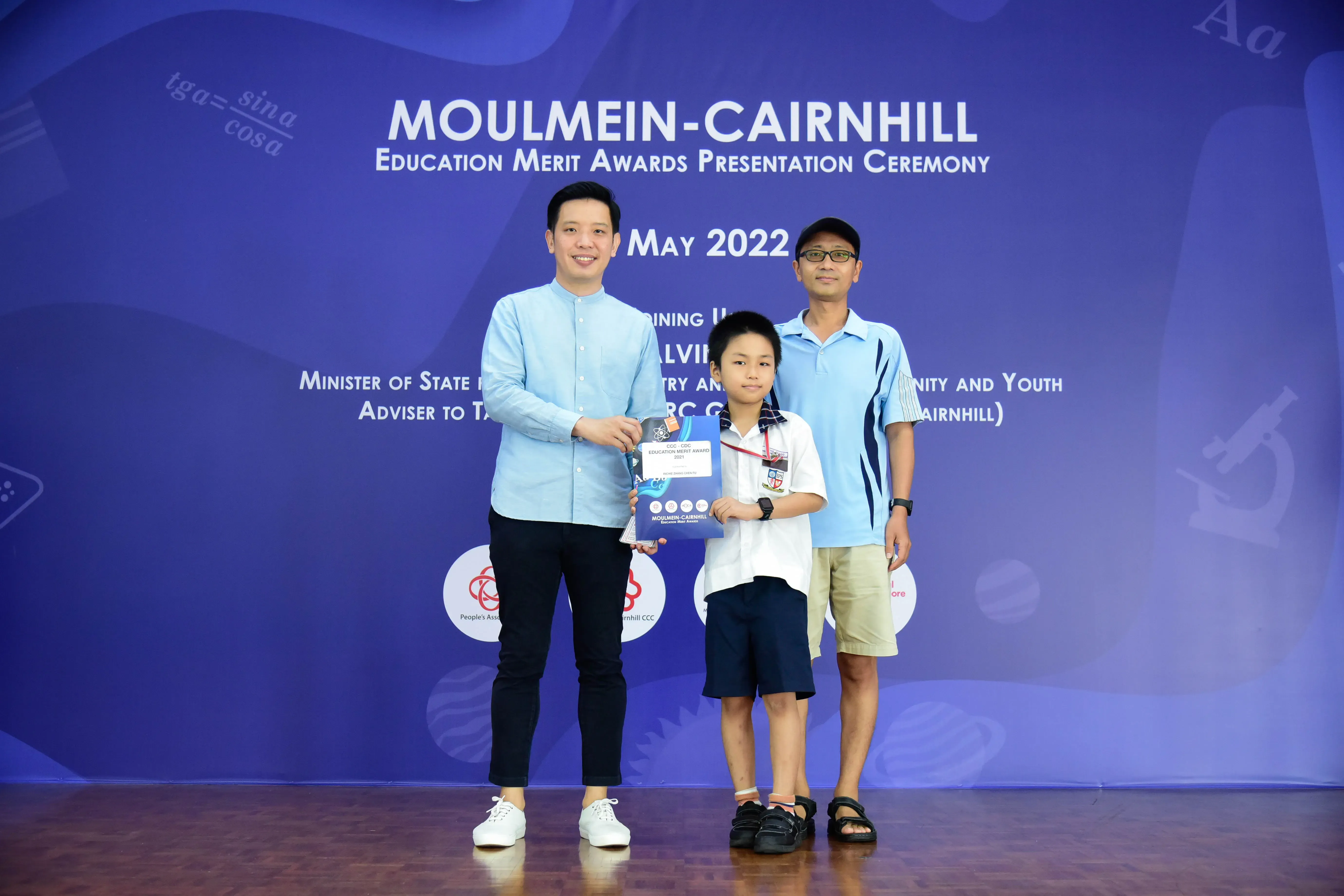 Three people stand on a stage at the Moulmein-Cairnhill Education Merit Awards Presentation Ceremony, May 2022.