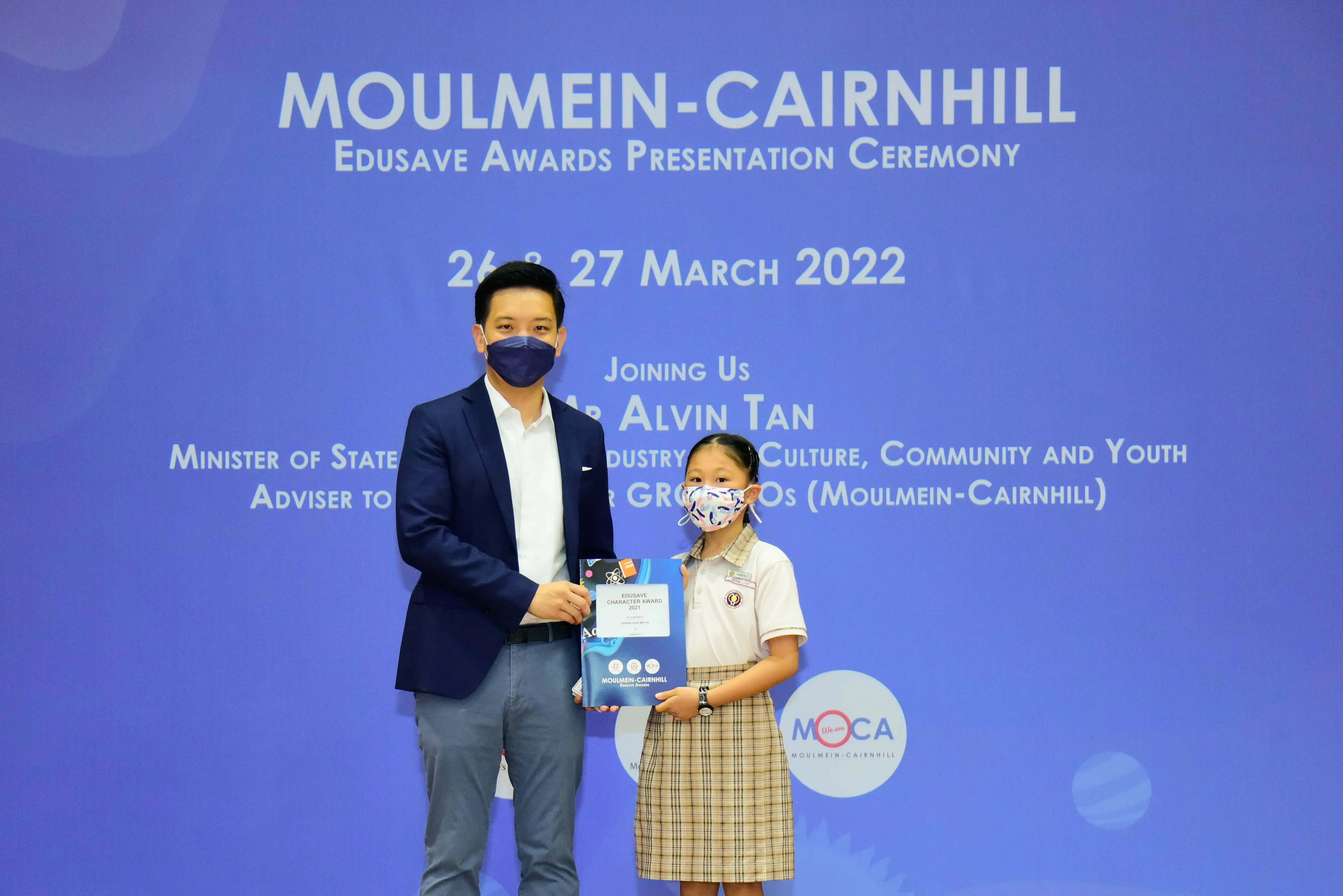 A masked man and young girl in school uniform hold an award at the Moulmein-Cairnhill Edusave Awards Ceremony 2022.