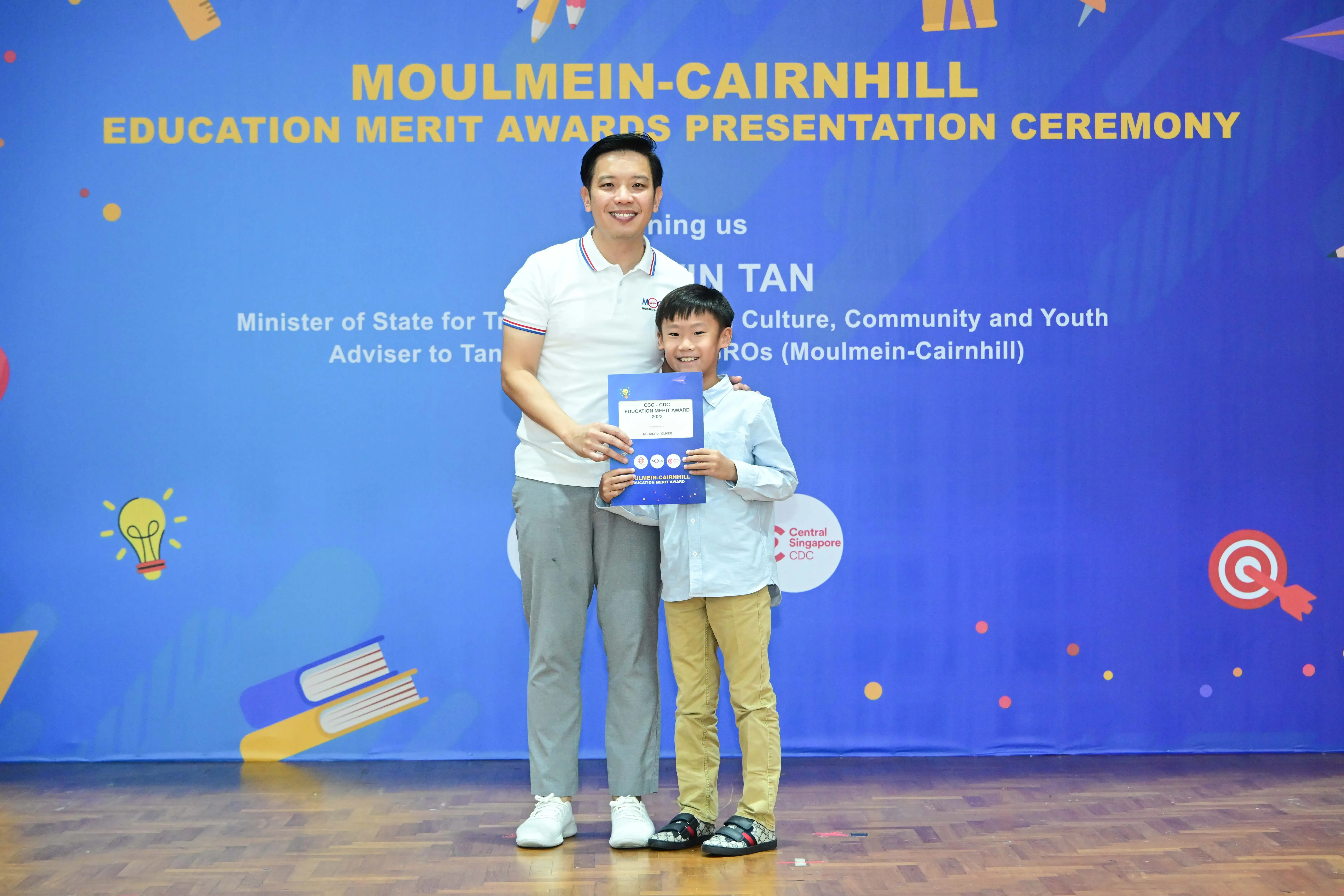 A man and a young boy holding a blue certificate at the Moulmein-Cairnhill Education Merit Awards ceremony.