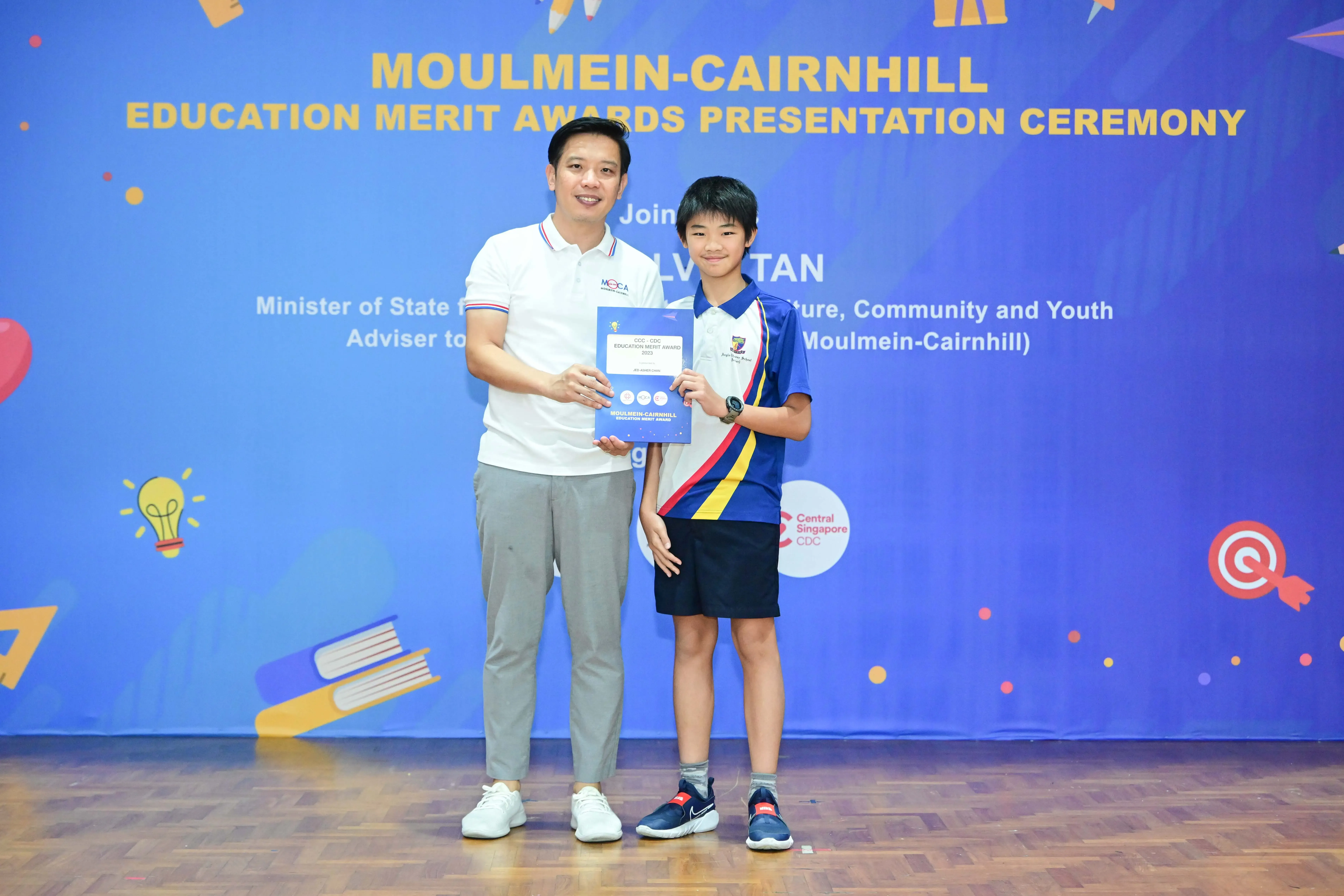An adult and a young student hold an award certificate at the Moulmein-Cairnhill Education Merit Awards Ceremony.