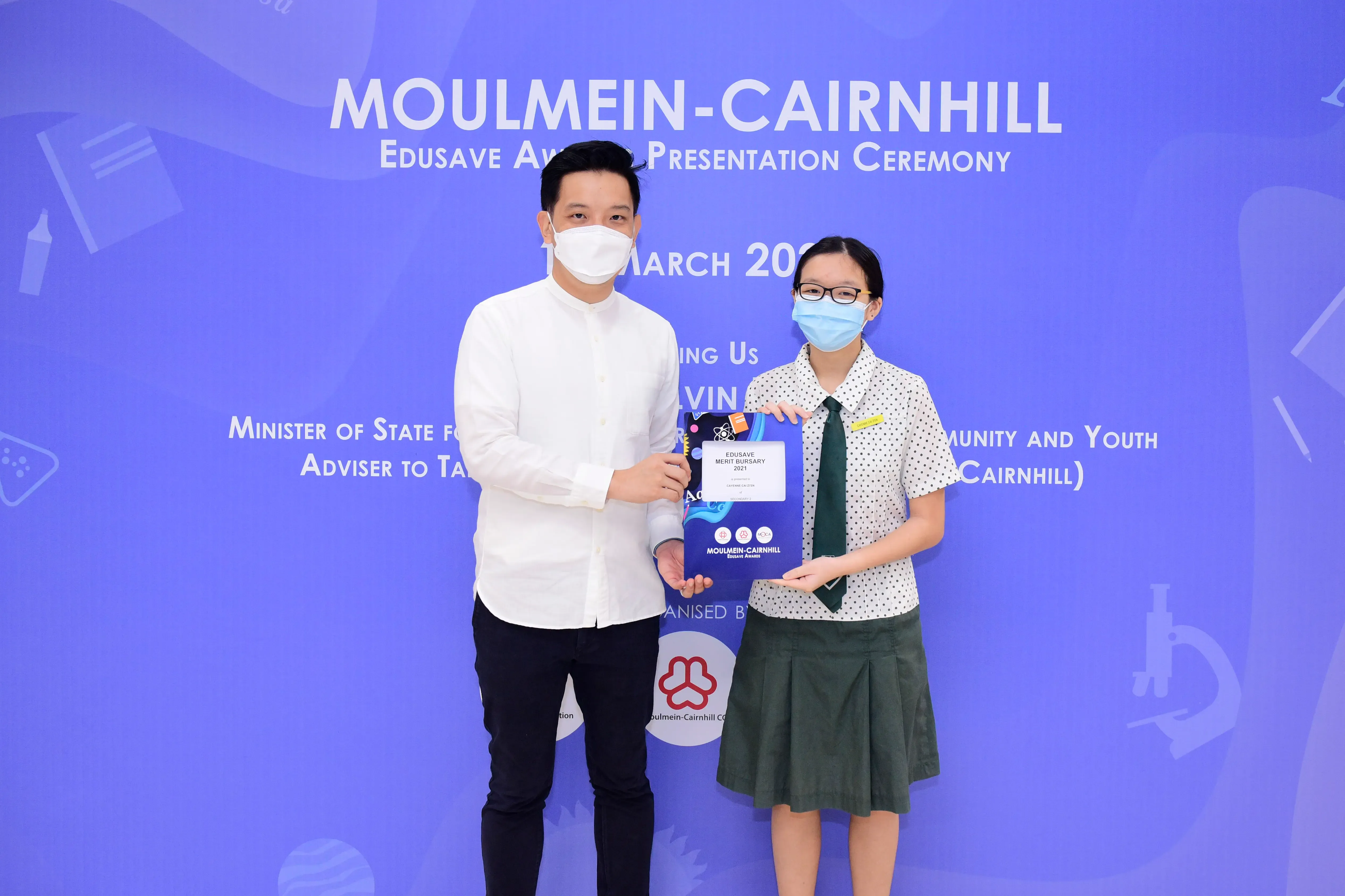 Two masked individuals hold an Edusave Merit Bursary award at the Moulmein-Cairnhill presentation ceremony.
