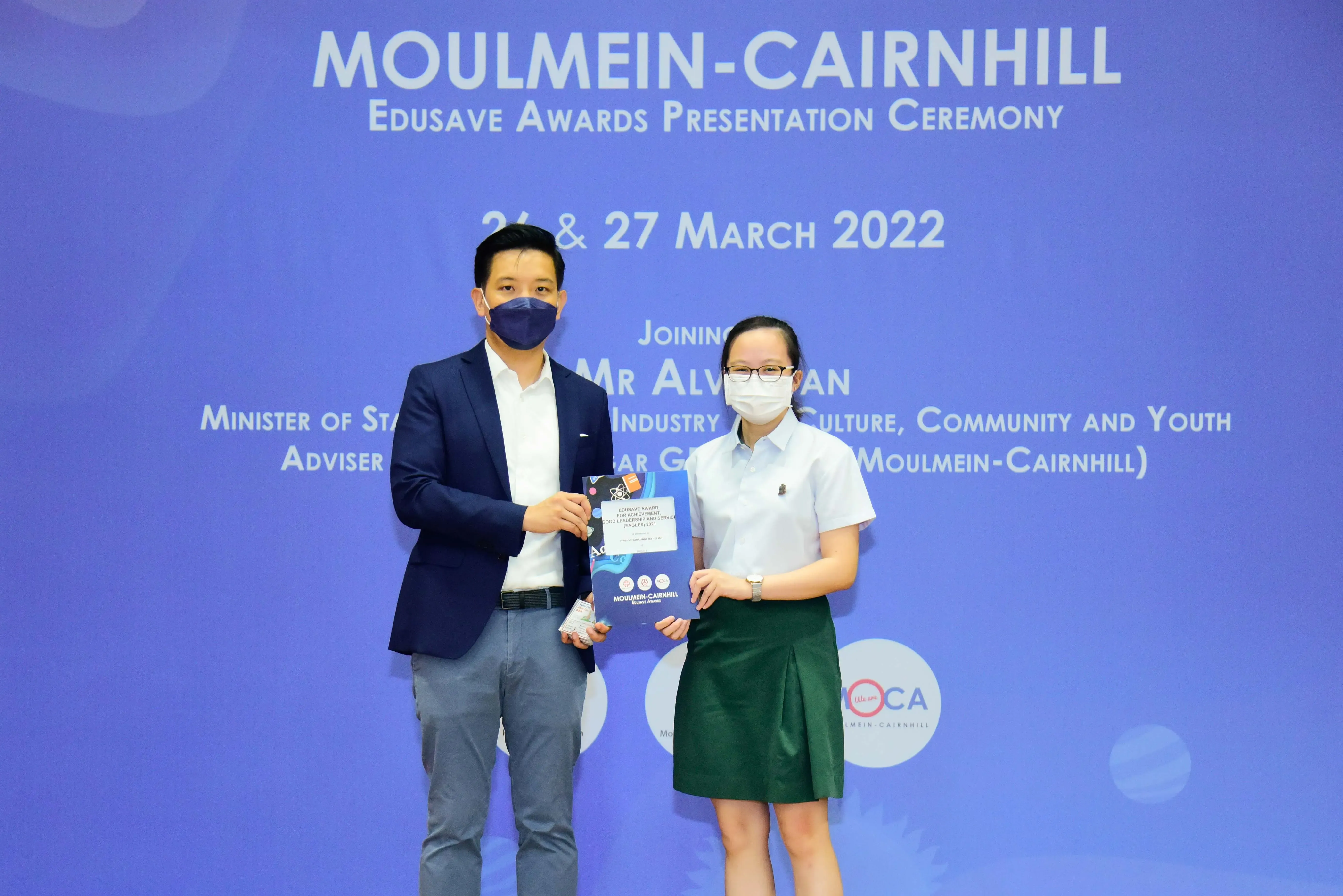 Two masked people hold an award booklet at the Moulmein-Cairnhill Edusave Awards Presentation Ceremony, March 2022.
