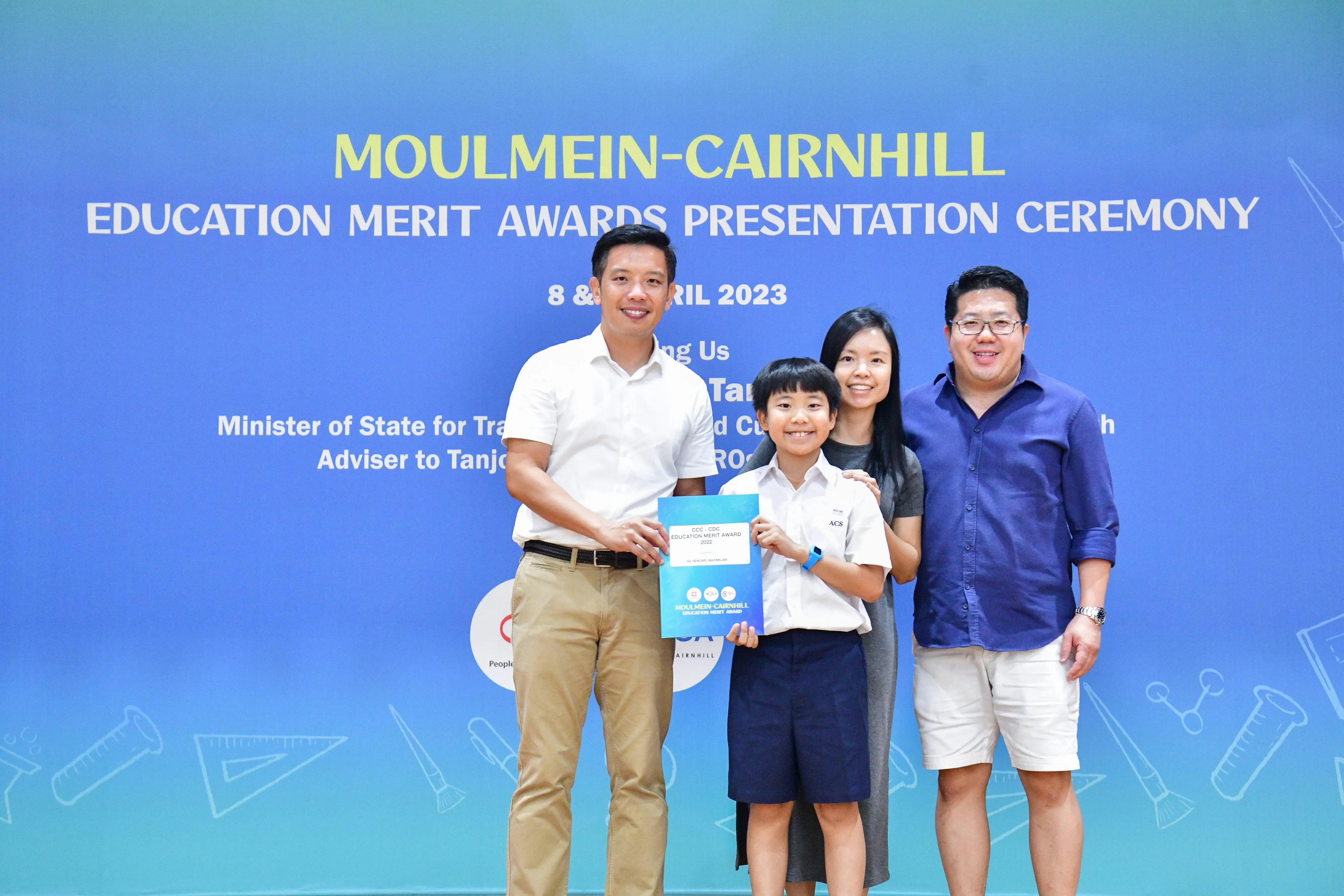 Four people pose before a Moulmein-Cairnhill Education Merit Awards Ceremony backdrop; a boy holds a certificate.