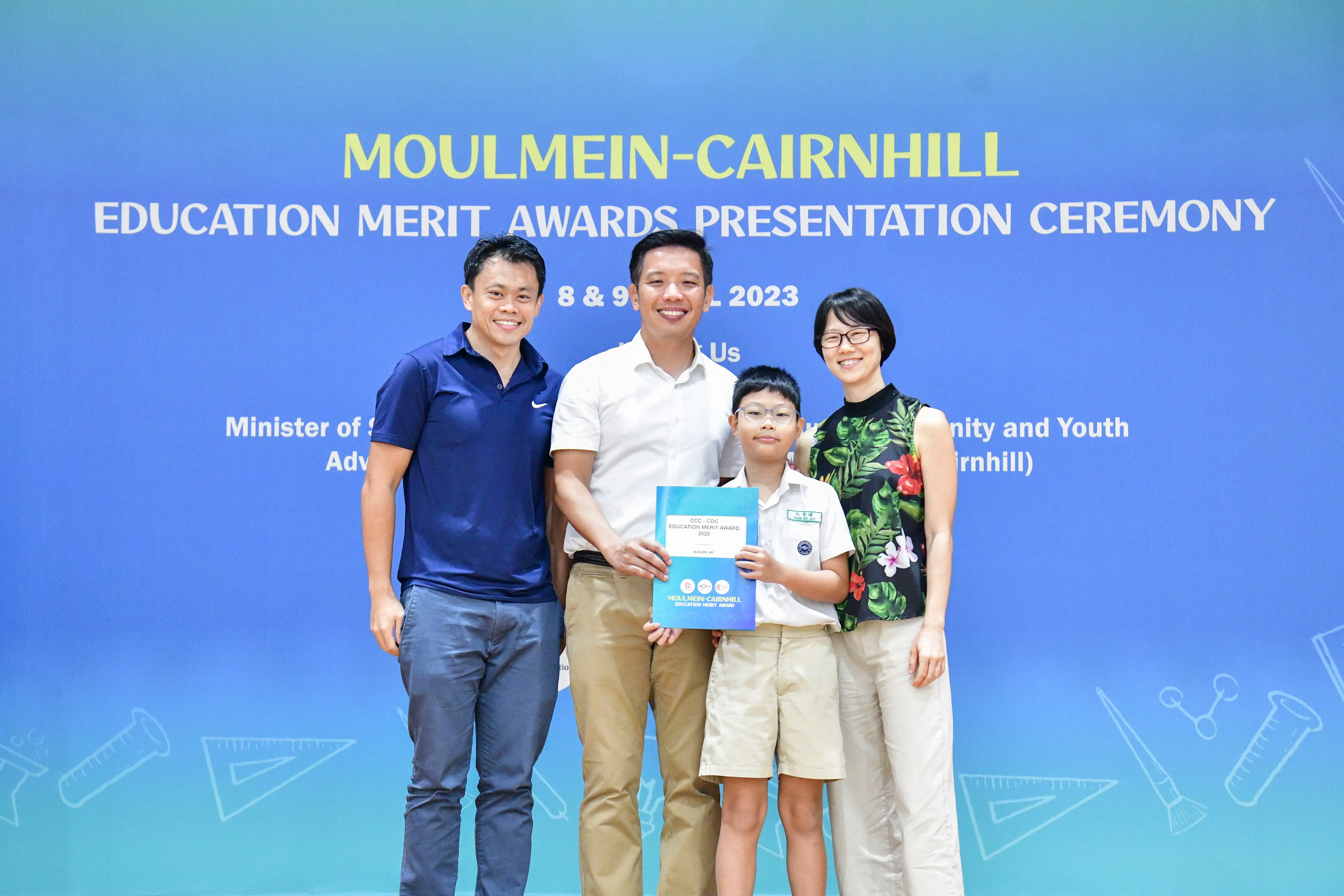 Four people pose before a Moulmein-Cairnhill Education Merit Awards Presentation Ceremony banner, a boy holding an award folder.
