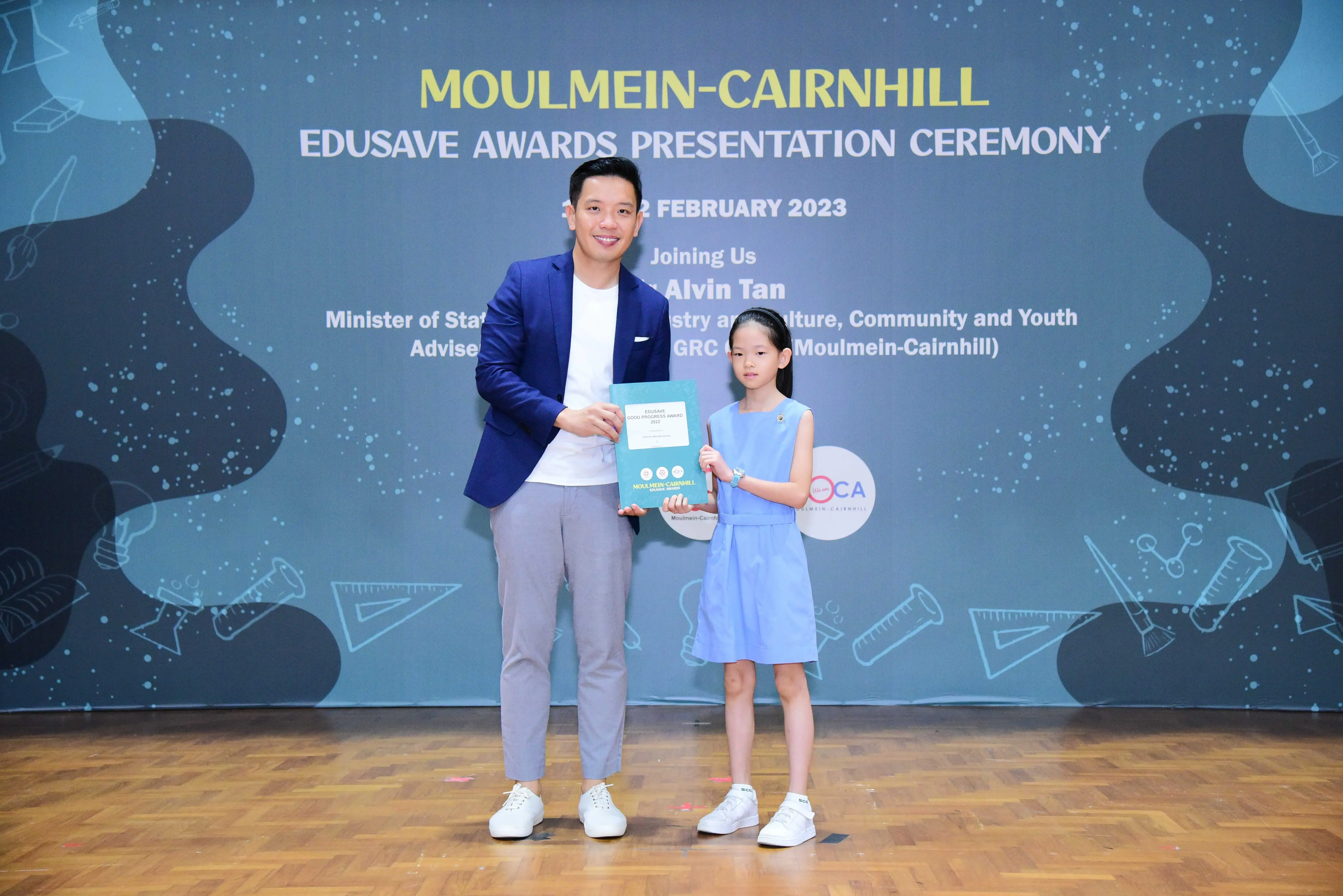 A man in a blue blazer and a young girl in a blue dress hold an award at the Moulmein-Cairnhill Edusave ceremony.