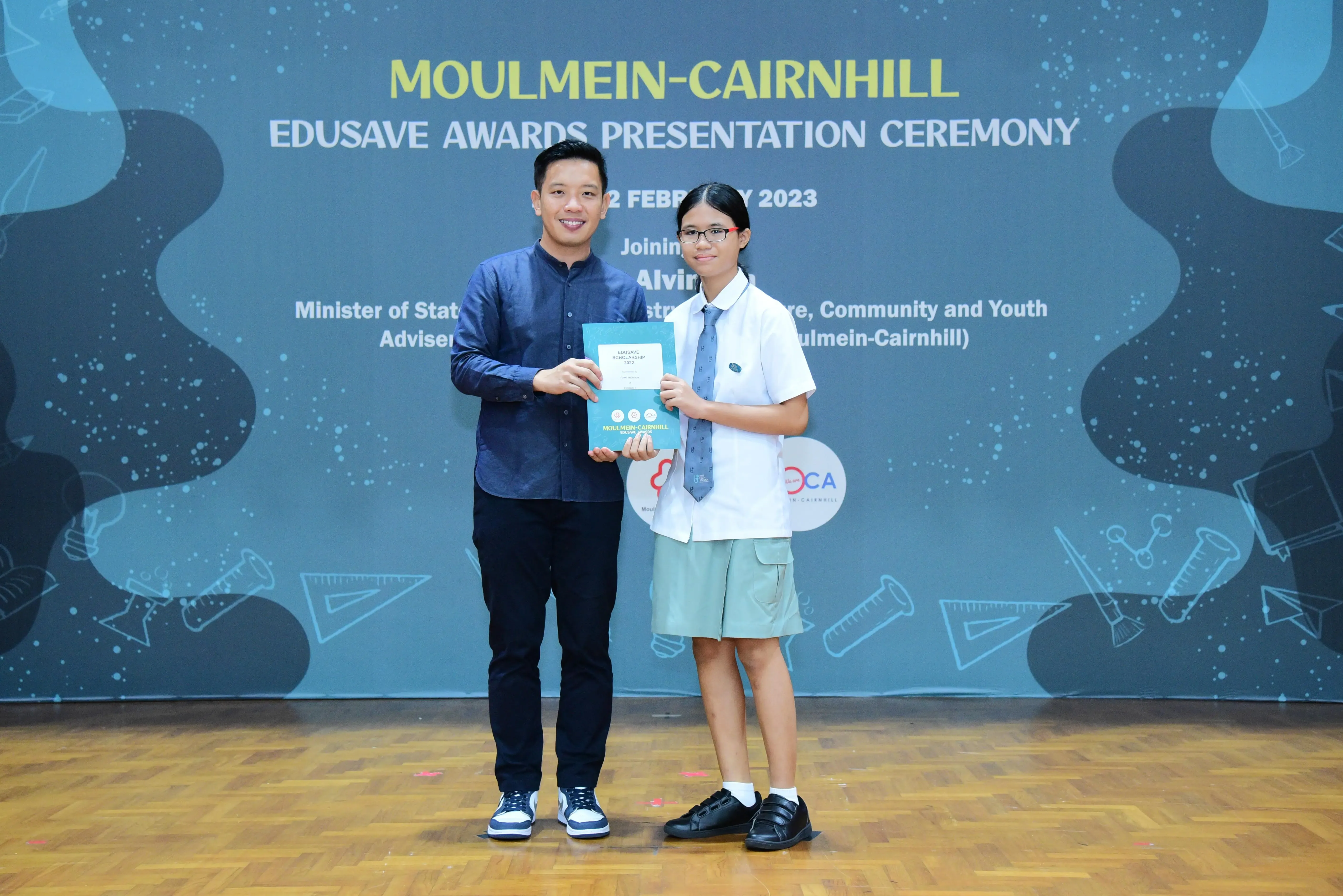 A man and a student in school uniform hold a certificate at the Moulmein-Cairnhill Edusave Awards Ceremony 2023.