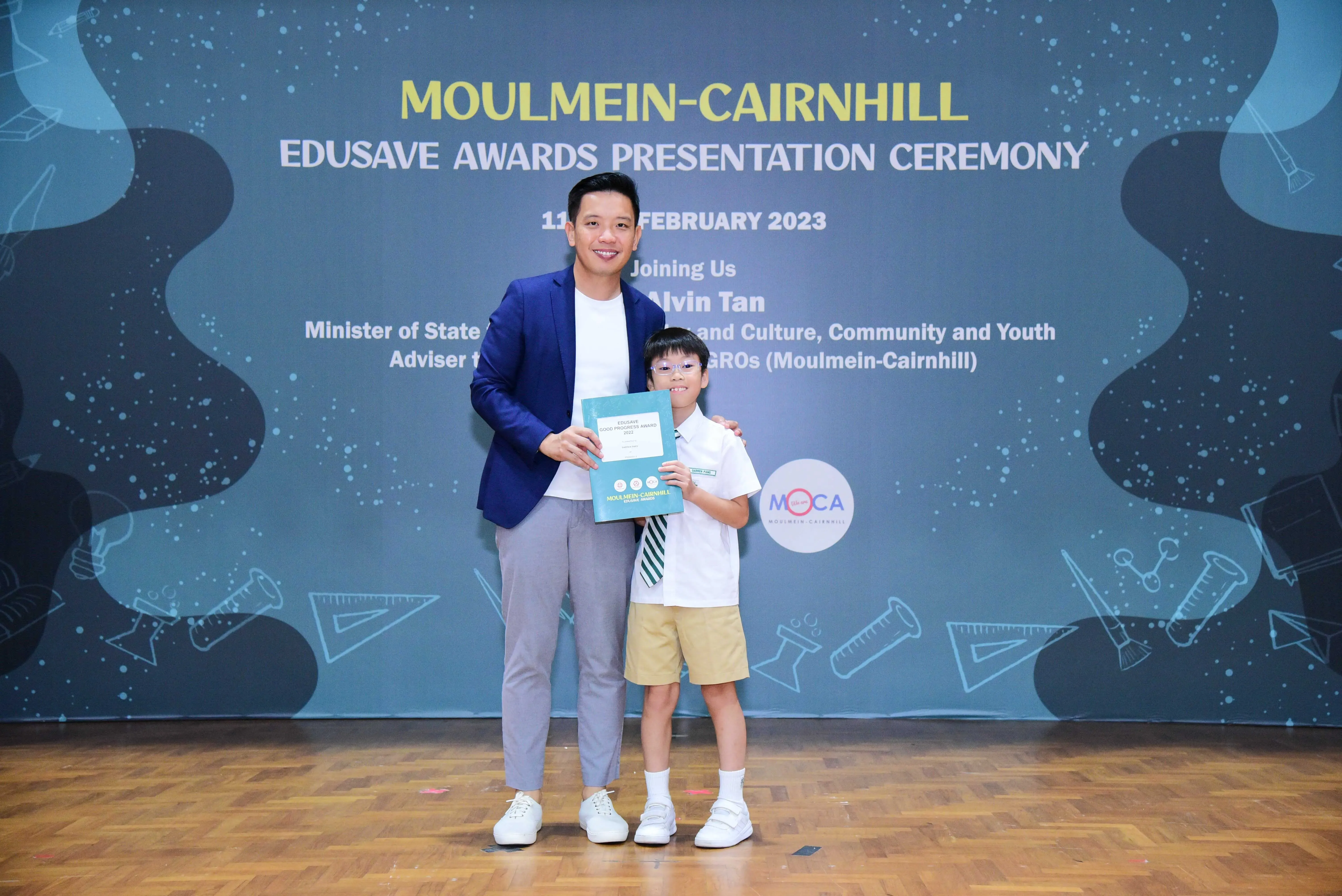 A man in a blue blazer and a young boy in school uniform hold an award at the Moulmein-Cairnhill Edusave Awards Ceremony 2023.