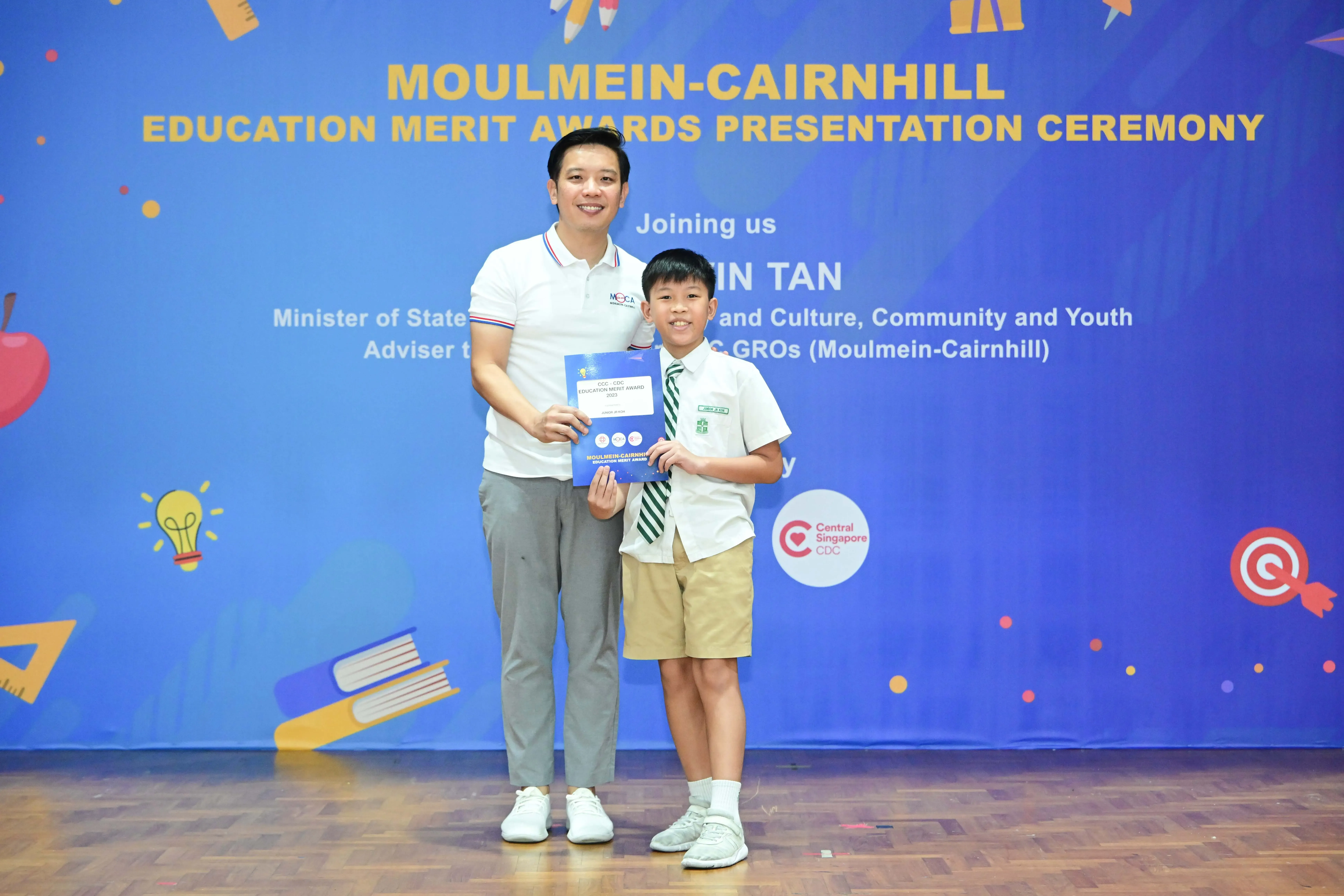 An adult and a young boy in school uniform hold an award certificate at the Moulmein-Cairnhill Education Merit Awards ceremony.