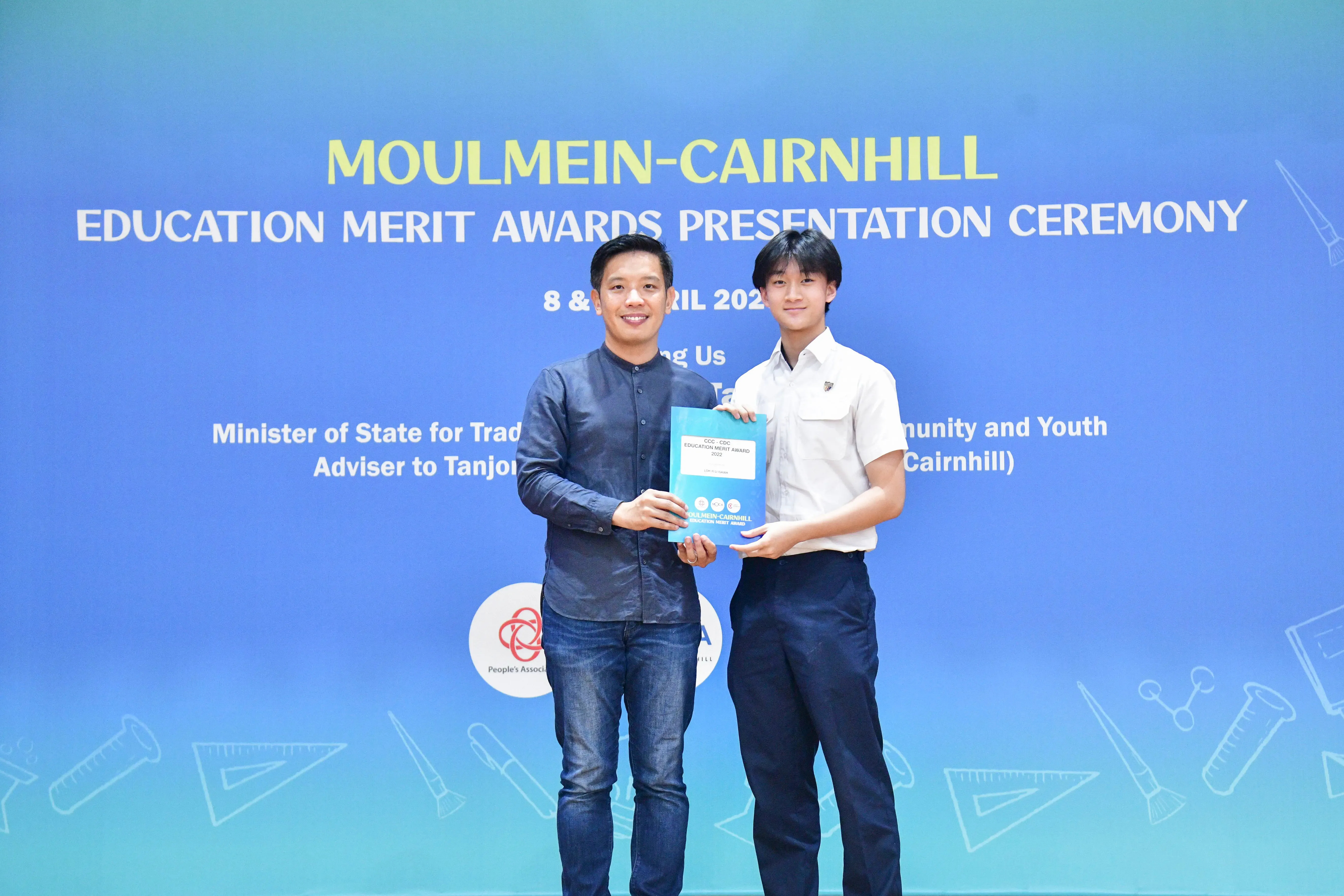 Two men hold a certificate together in front of a Moulmein-Cairnhill Education Merit Awards Presentation Ceremony banner.