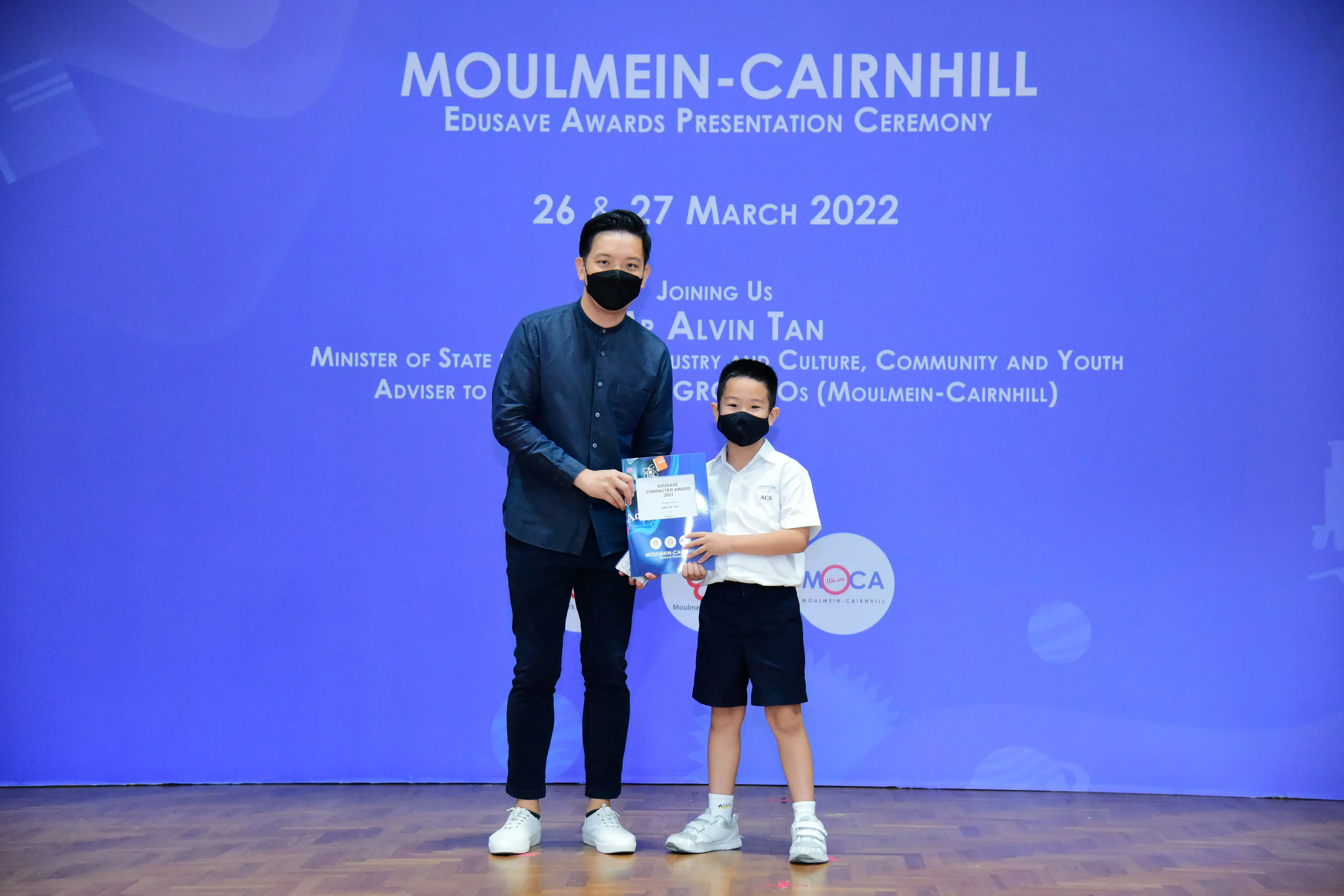 An adult and a young boy in masks hold a certificate at the Moulmein-Cairnhill Edusave Awards Ceremony, March 2022.