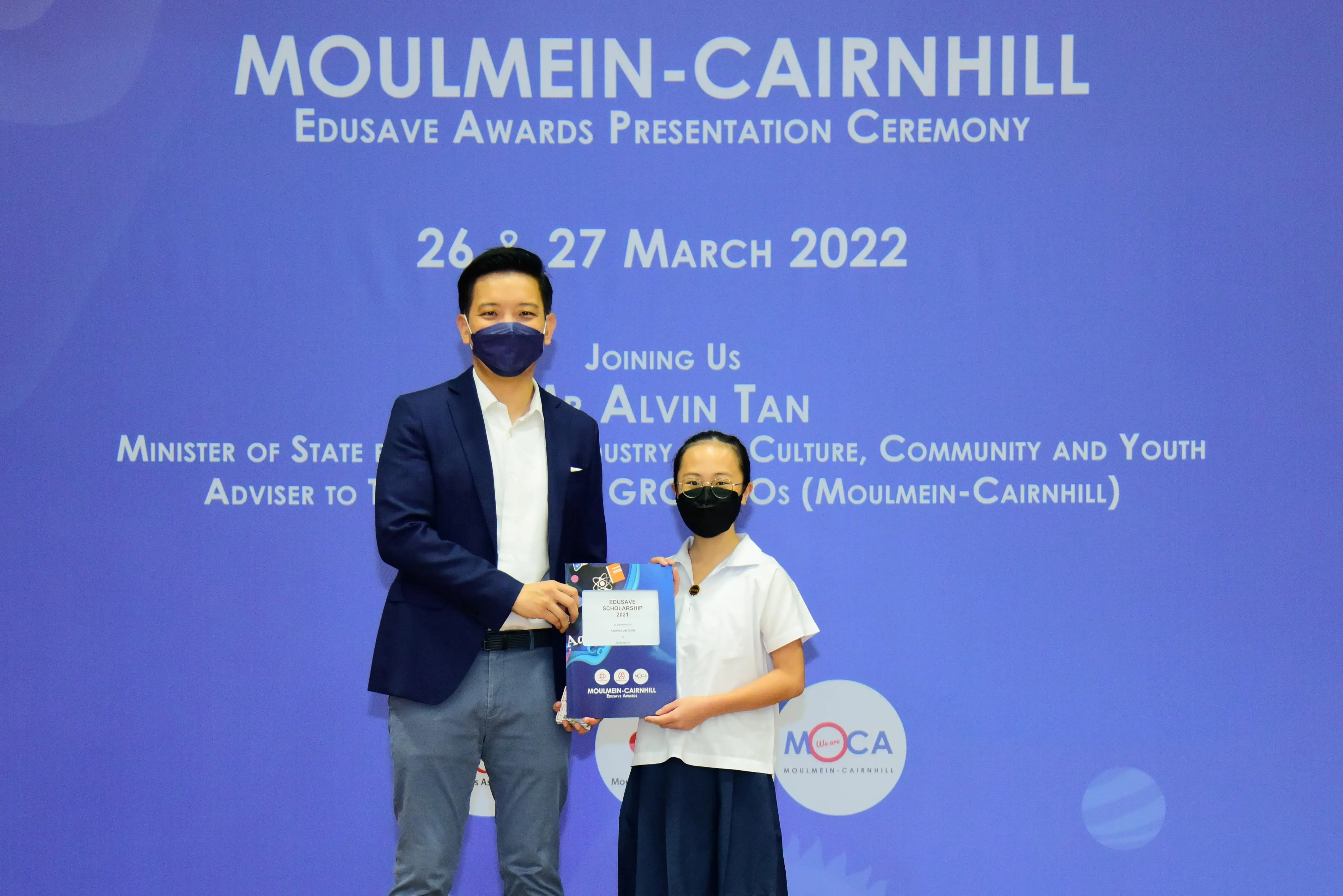 Two masked individuals hold an Edusave Scholarship certificate at the Moulmein-Cairnhill Awards Ceremony, March 2022.