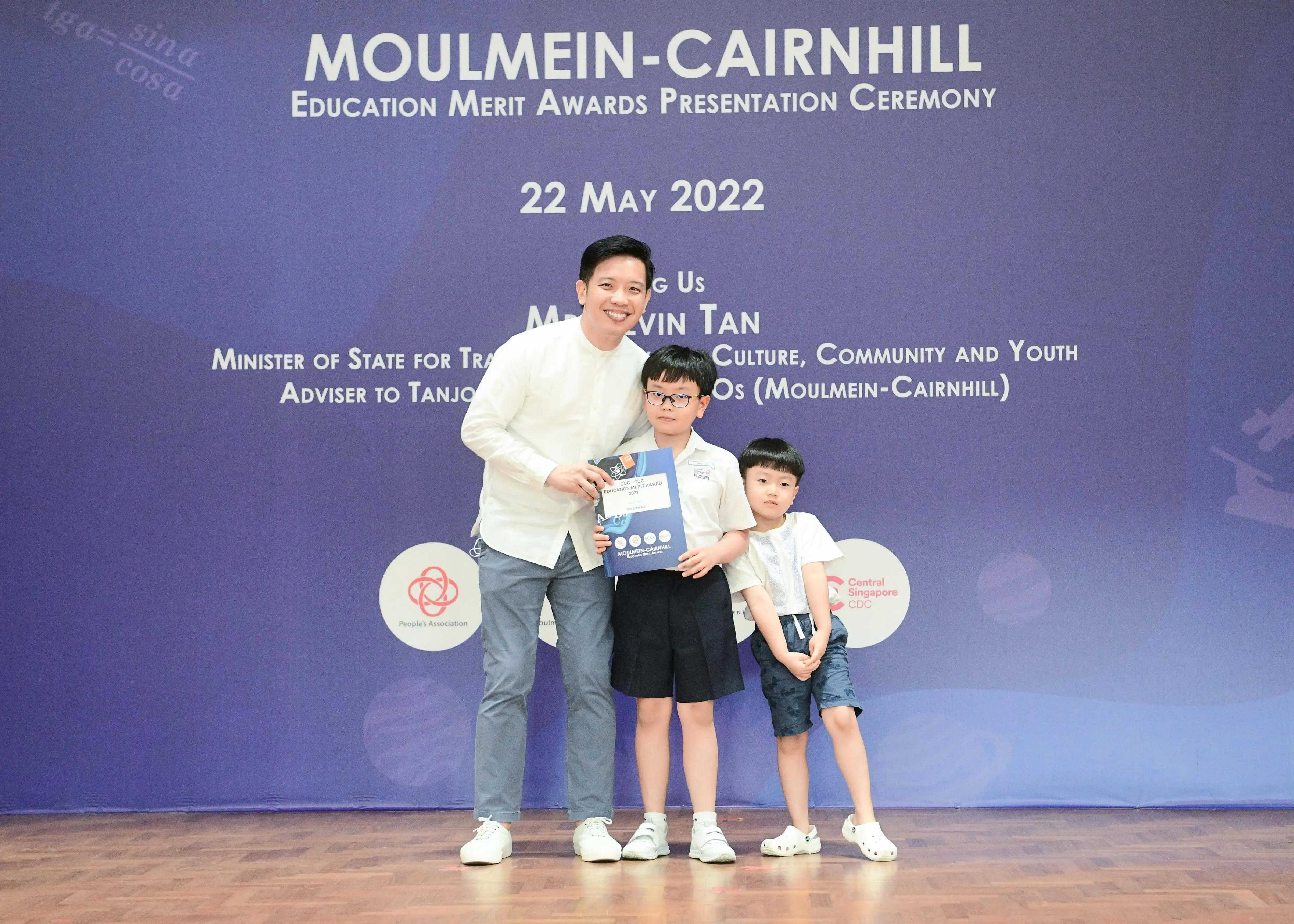 Man and two young boys pose holding an award booklet at the Moulmein-Cairnhill Education Merit Awards Ceremony, May 2022.