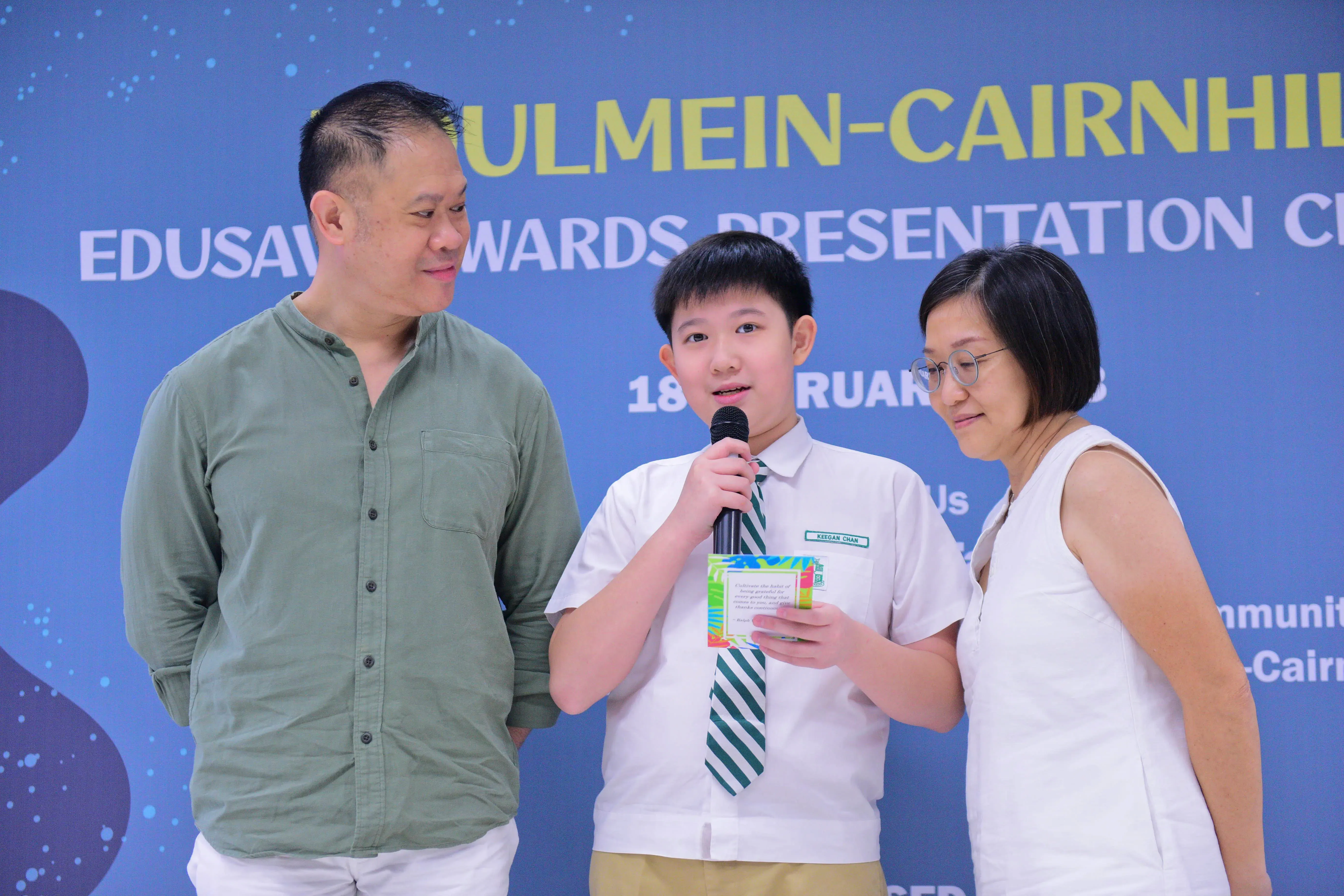 A boy in school uniform speaks into a microphone holding a card, flanked by two adults at an Edusave Awards ceremony.