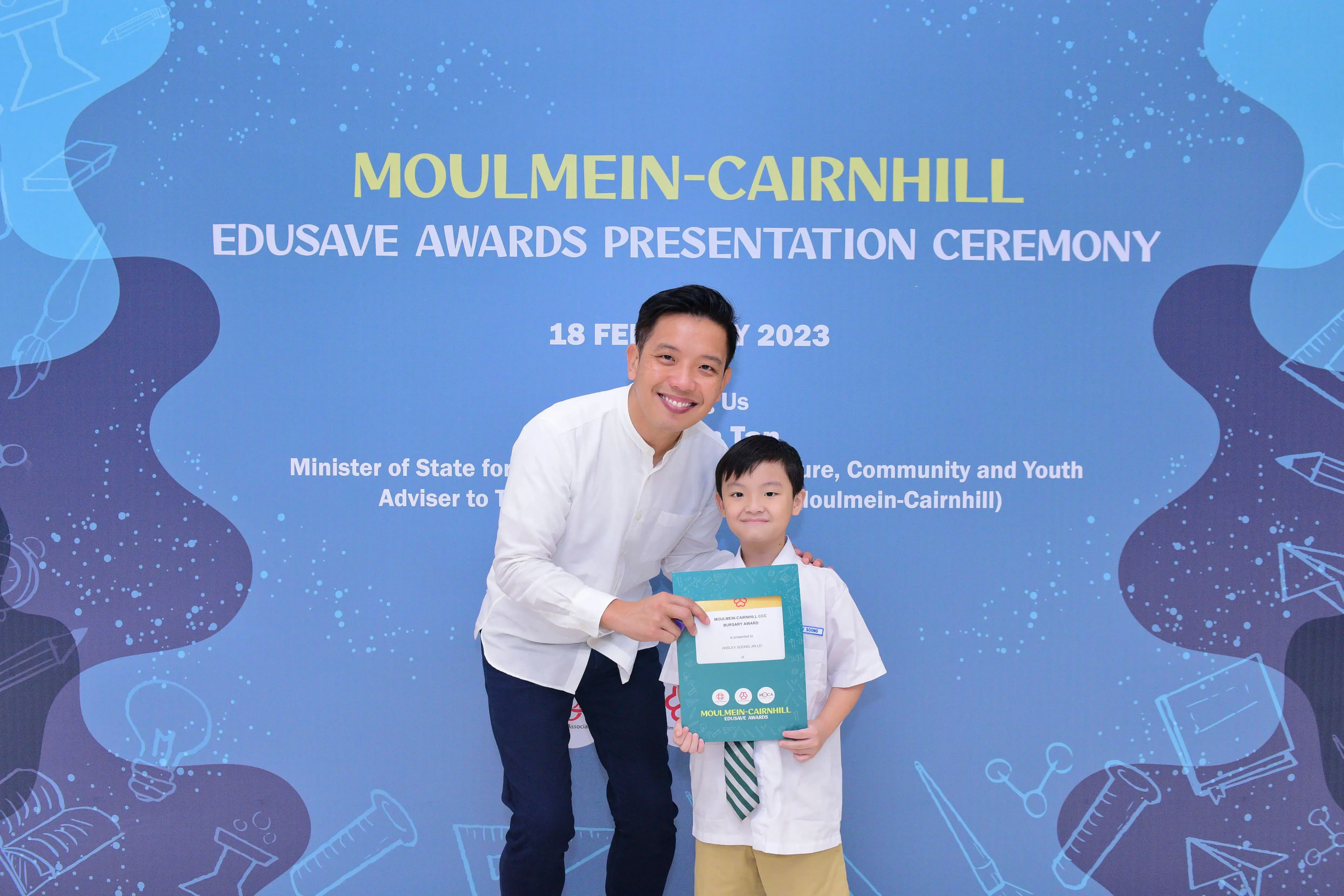 A man and a young boy hold a certificate in front of a Moulmein-Cairnhill Edusave Awards Ceremony banner.