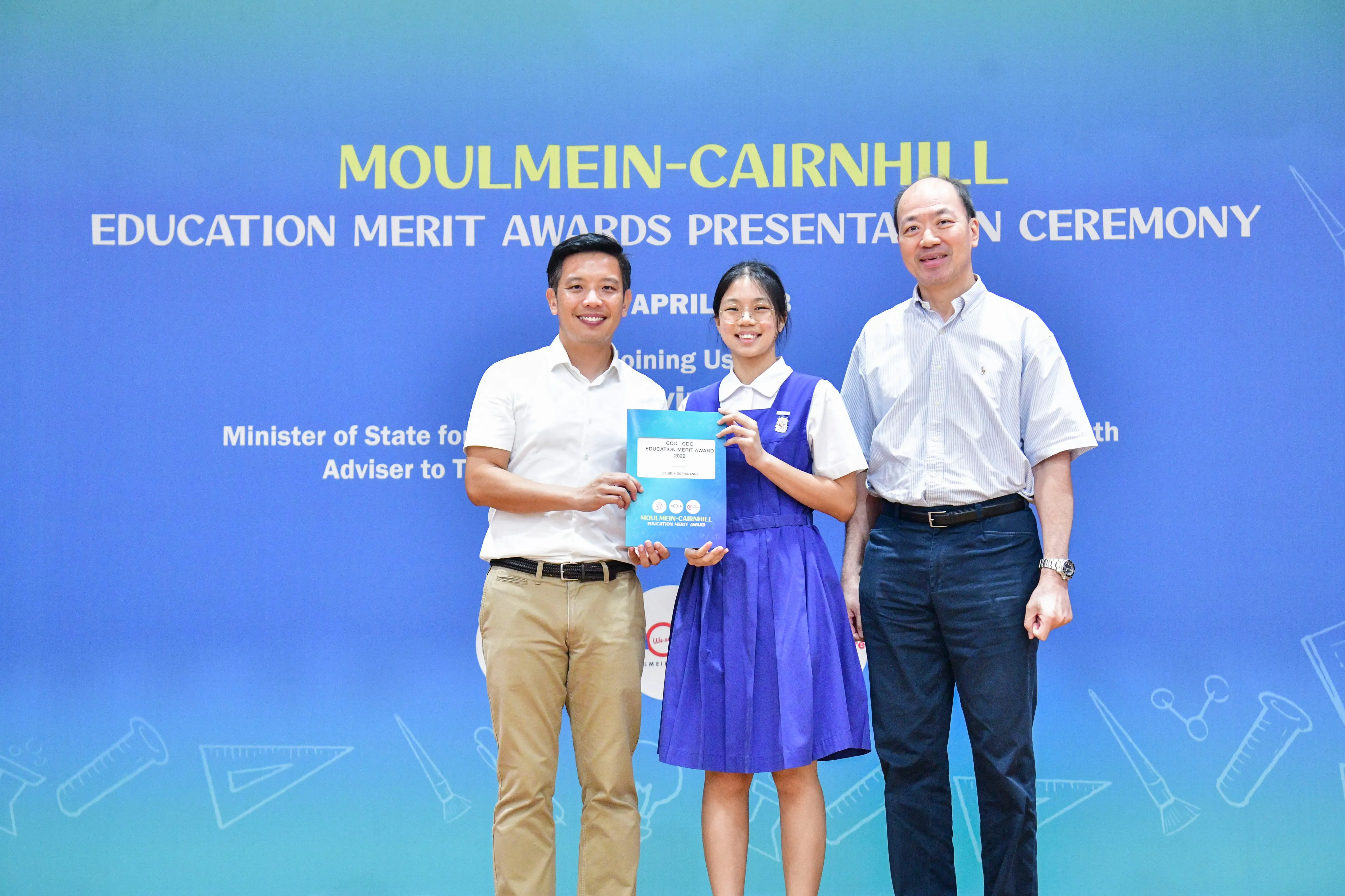Three people pose at the Moulmein-Cairnhill Education Merit Awards Presentation Ceremony, with a student holding an award.