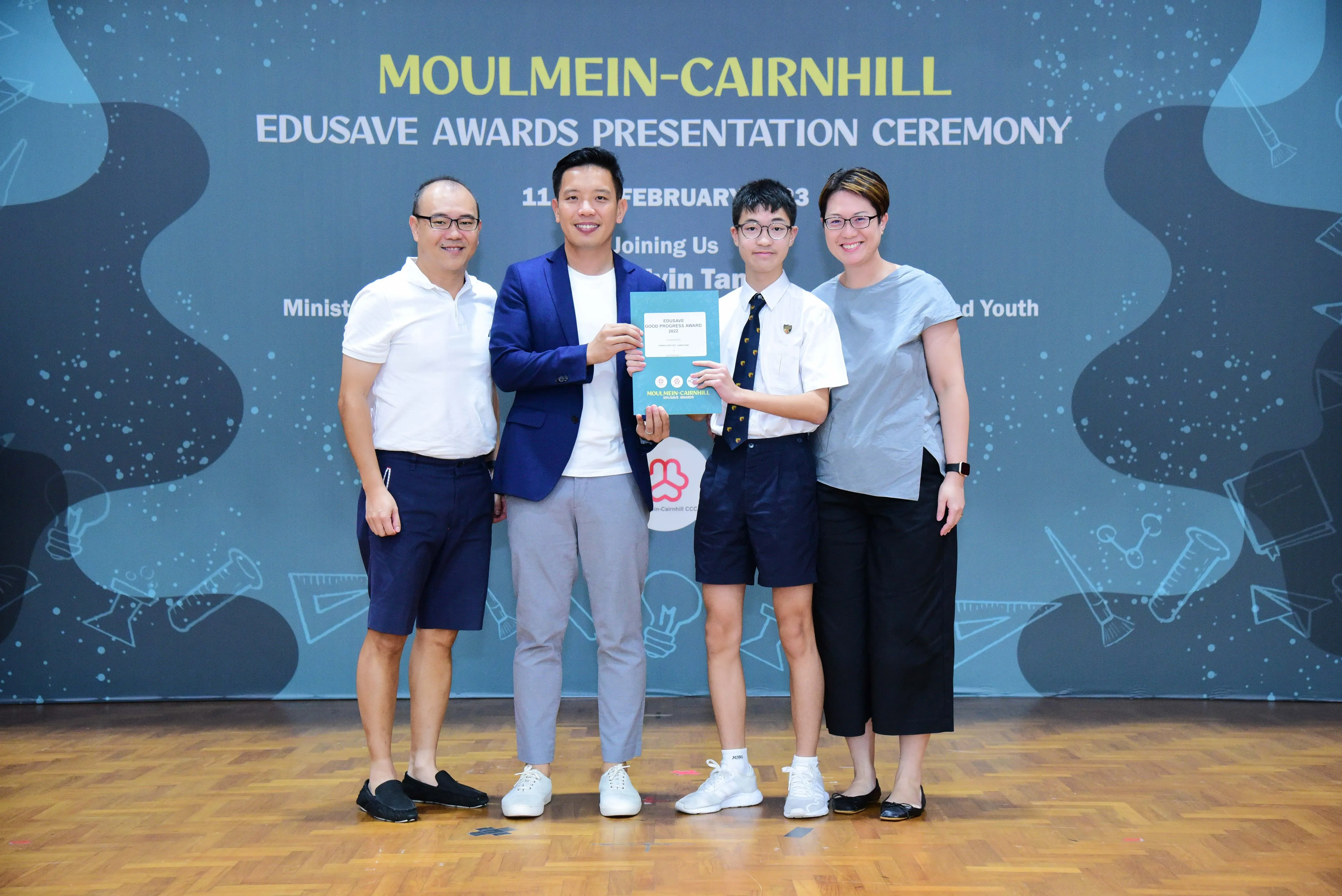 Four people pose holding an award certificate at the Moulmein-Cairnhill Edusave Awards Presentation Ceremony backdrop.