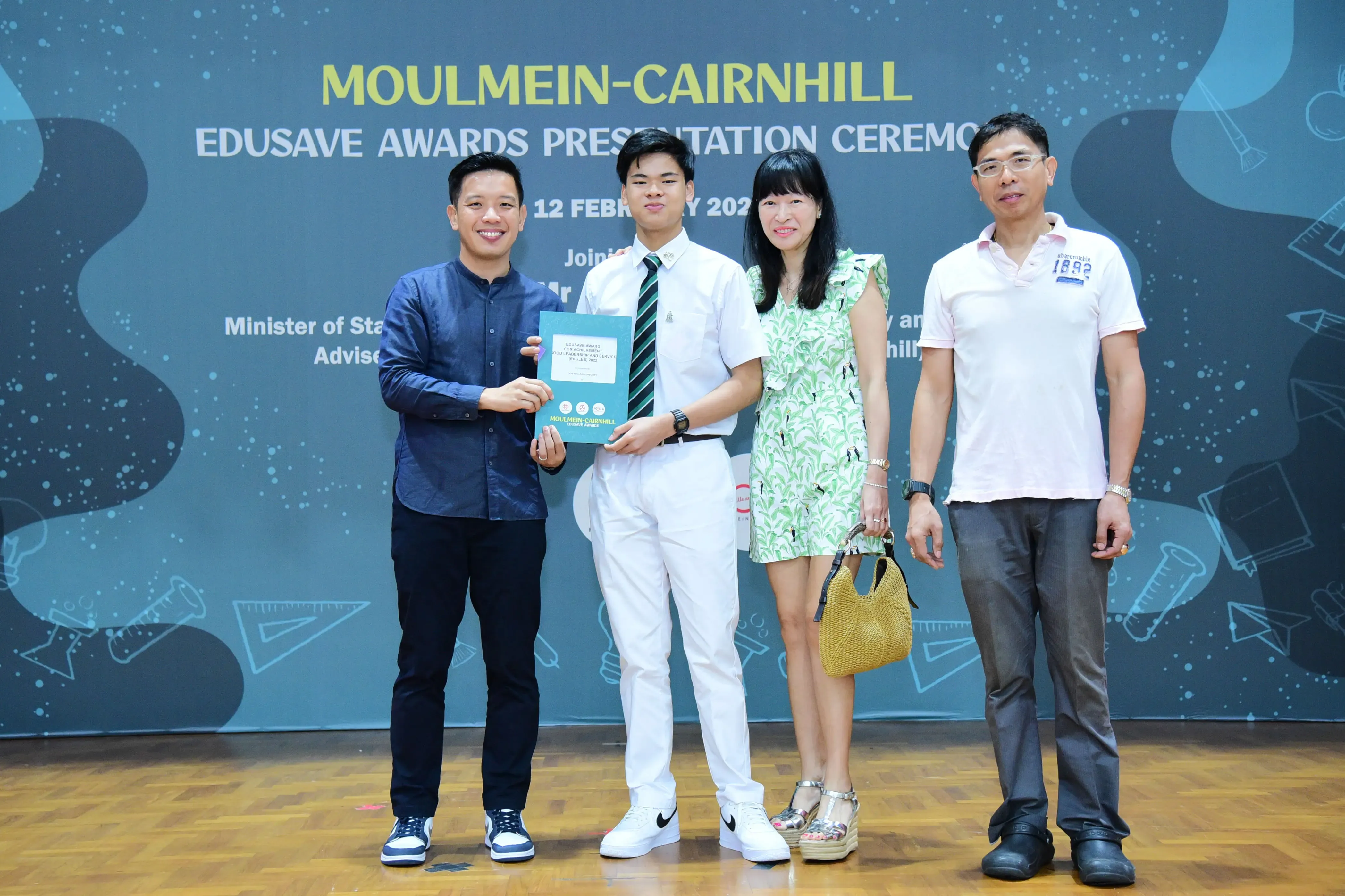 Four people pose on stage at the Moulmein-Cairnhill Edusave Awards Presentation Ceremony, holding a certificate.