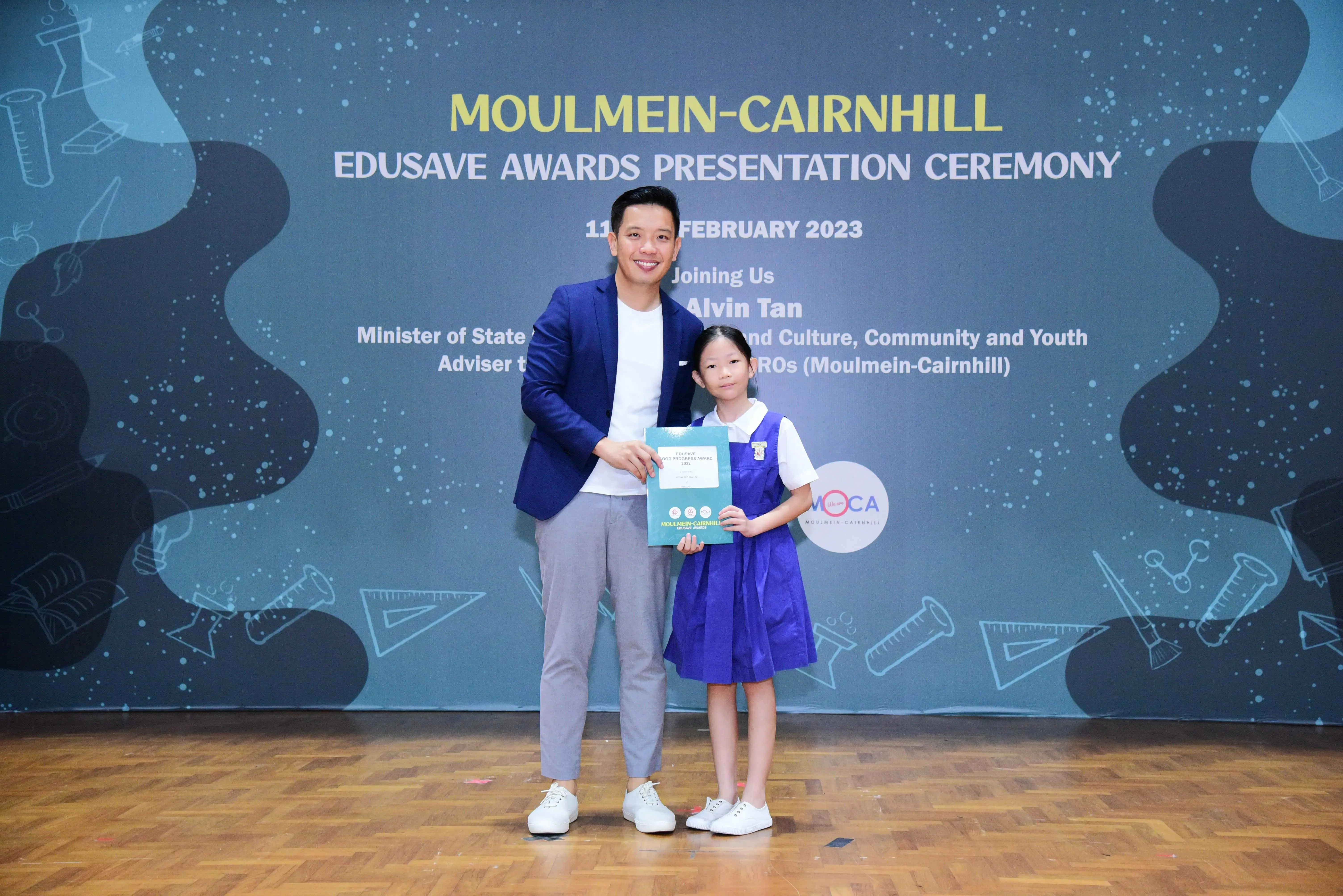 A man in a blue blazer and a young girl in a blue uniform hold a certificate at the Moulmein-Cairnhill Edusave Awards 2023.