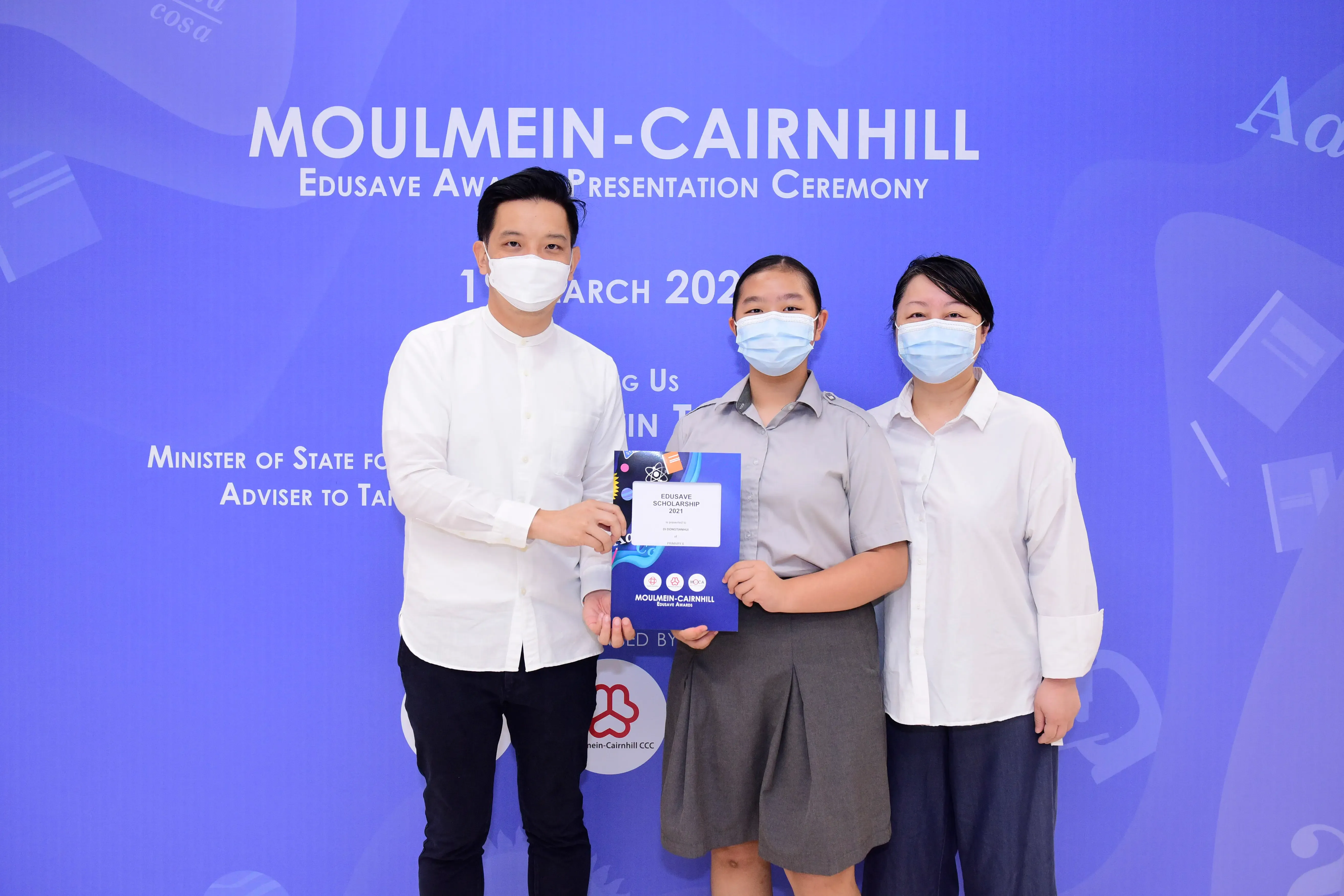 Three masked people pose at the Moulmein-Cairnhill Edusave Awards Presentation Ceremony, holding a scholarship certificate.